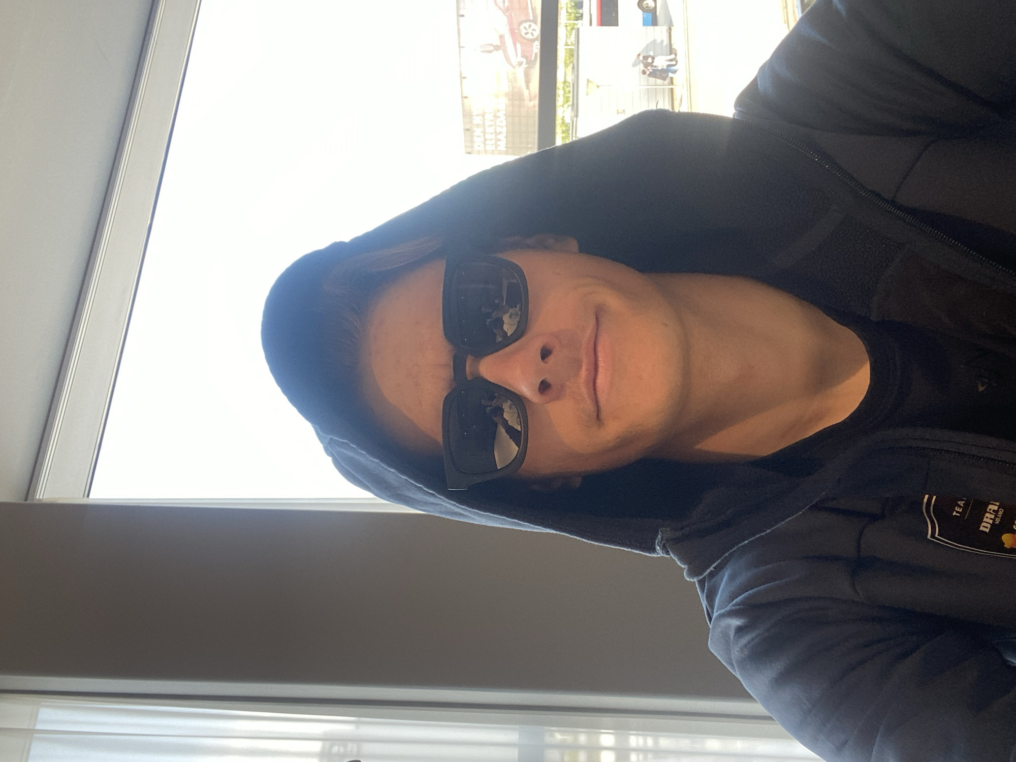 A selfie-style, rotated image shows a person wearing a dark hoodie and dark sunglasses. A window is visible in the background, showing a bright outdoor scene. Reflected in the sunglasses are images of buildings and trees, suggesting an urban setting. A billboard partially visible outside the window features an image of a car and text in what appears to be Danish. The person is indoors, next to the window. The logo on the hoodie reads “DRAI MILANO” and “TEA”. The location is likely in Kastrup, Denmark, based on the billboard signage.