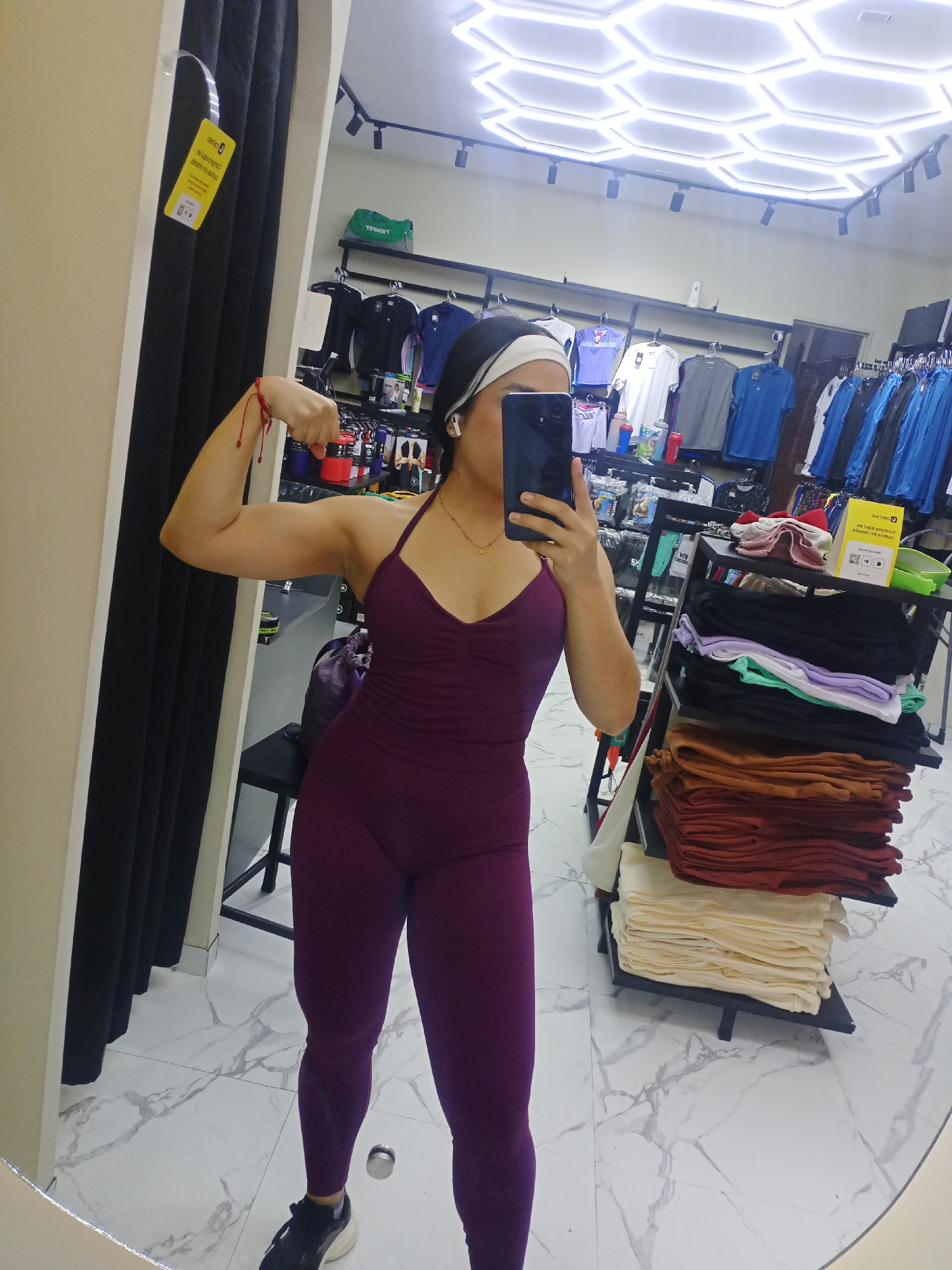 A person is captured in a mirror selfie taken inside a retail store, likely an athletic or casual wear shop in Valle de La Pascua, Venezuela. The individual, appearing to be female, is flexing their right bicep while holding a dark-colored smartphone to take the picture, partially obscuring their face. They are wearing a form-fitting, dark purple (plum) full-body jumpsuit or unitard with a v-neck halter top and leggings, paired with black and white athletic shoes. A small gold heart pendant necklace is visible, as are white wireless earbuds and a white headband. A red string bracelet is tied around the right wrist.
The setting appears to be a changing room area or a section of the store. To the left, a black changing room curtain or partition is visible, from which a yellow rectangular tag hangs. To the right, various clothing racks display t-shirts, shorts, and other apparel. A green duffel bag with the mirrored text "TENUT" sits on a shelf above the racks. Below, a black shelving unit neatly displays stacks of folded garments in various colors including black, light green, purple, orange, red, and cream/off-white. Another yellow tag, similar to the one on the left, is positioned on this shelf. The floor is covered with white marble-patterned tiles, and the ceiling features modern hexagonal LED light panels along with track lighting.
Visible text on the yellow tags includes the brand name "Perfect" and partial Spanish text that appears to read "no logo commo..." (likely "no logo común" or "no logo common") and "no usar en público" (meaning "do not use in public"), possibly indicating sample items or internal store markings. The overall scene suggests a vibrant, well-lit retail environment.