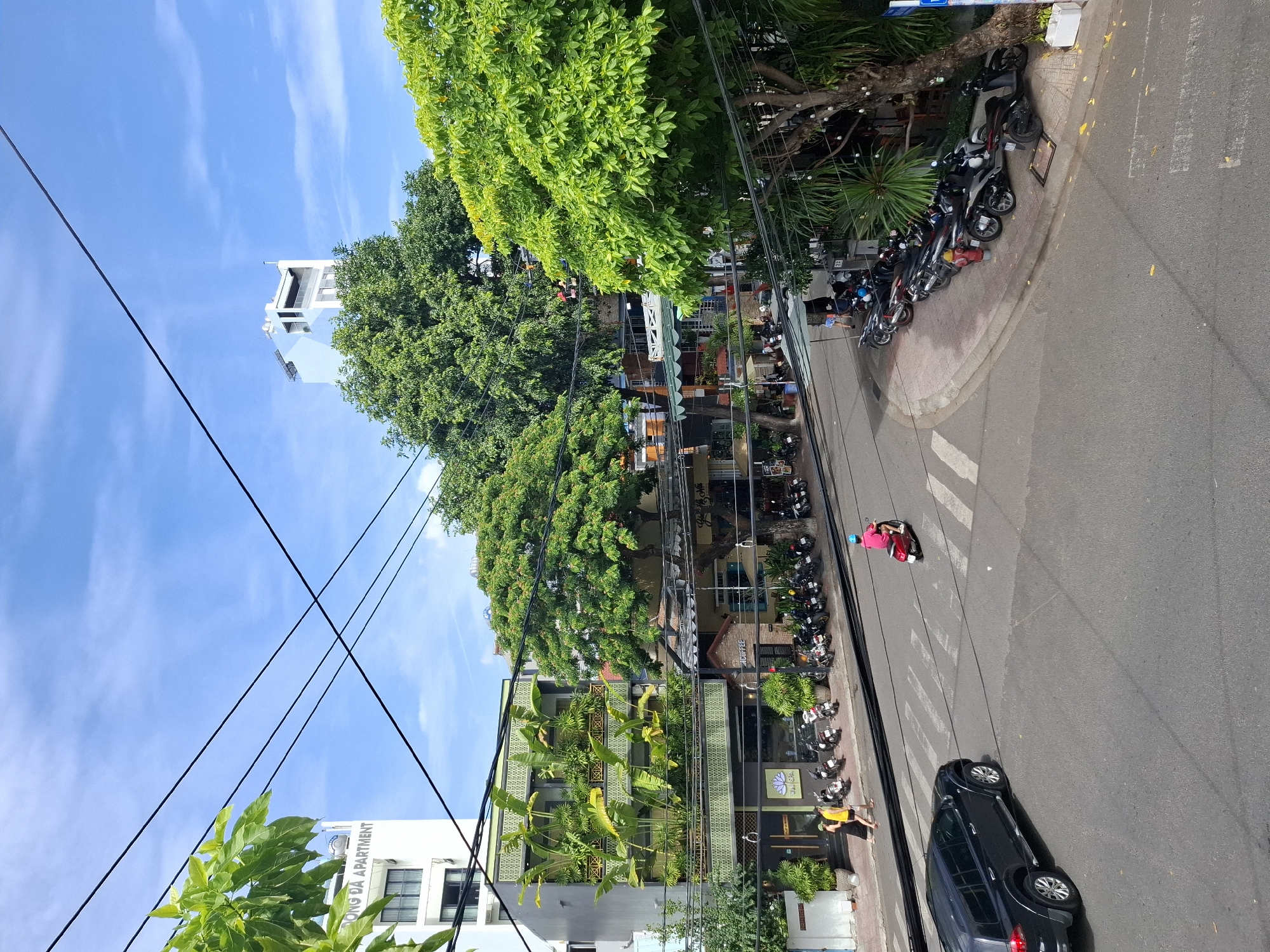 In Nha Trang, Vietnam, a street scene unfolds under a bright, partly cloudy blue sky. A black SUV is driving across a pedestrian crossing on the right side of the road. Further up the street, a person on a pink scooter is also crossing. On the left sidewalk, two individuals, one in a yellow top and the other in black, walk towards a building with a lotus flower logo. The sidewalk is lined with parked motorcycles. The buildings lining the street feature a mix of modern architecture and lush greenery, with several balconies adorned with plants. A tall, white building with "BONG DA APARTMENT" visible on its side stands in the background. Overhead, a network of power lines crisscrosses the sky.