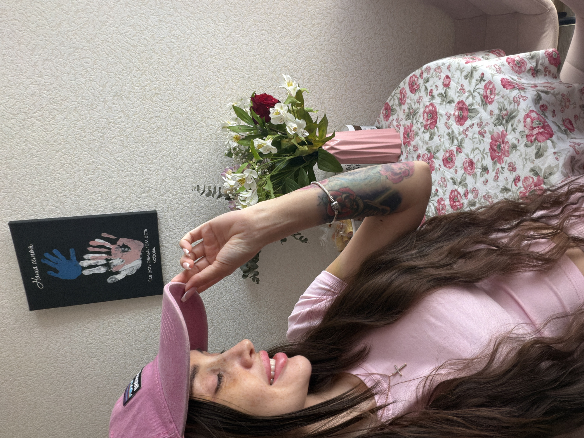 A woman lies on a floral patterned chair or bed wearing a pink shirt and baseball cap. She is smiling and appears relaxed. On the wall beside her is a black book with colorful handprints and Russian text that translates to "Our family, Where there is family, there is love". Beside the book is a pink vase containing a bouquet of flowers including a red rose. The environment appears to be indoors. There is no information to indicate this location is in Saint Petersburg, Russia.