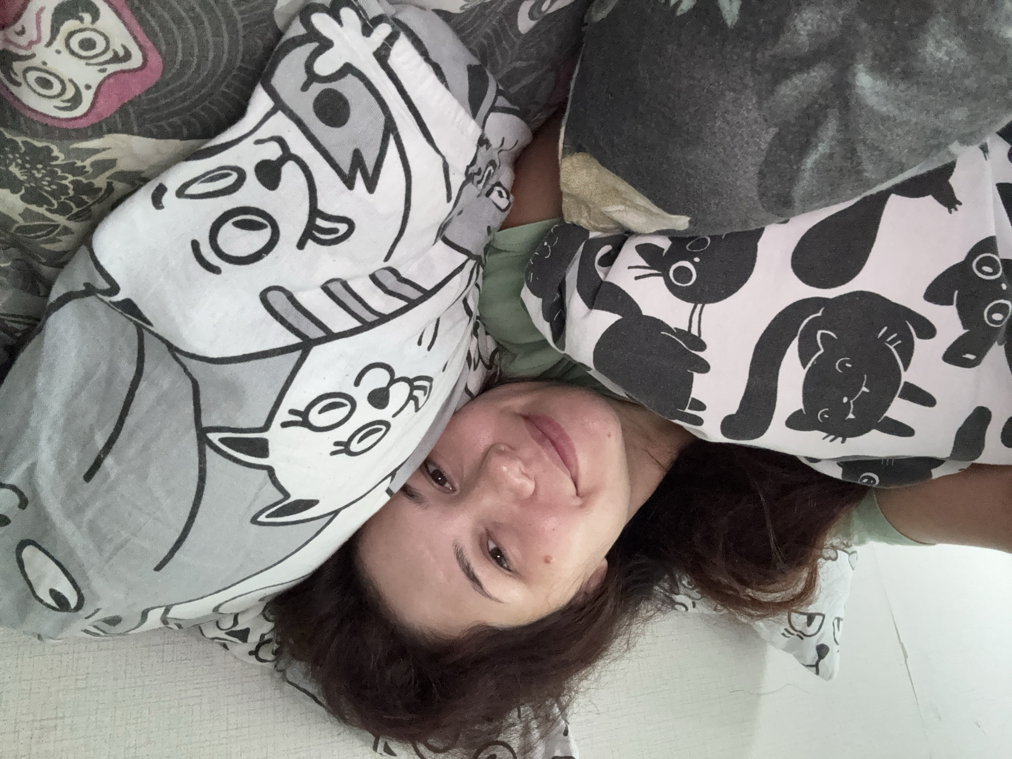 A woman with dark, curly hair is lying in bed, partially covered by pillows and blankets. She is looking at the camera with a slight smile. The pillows and blankets have cartoon cat patterns. The background appears to be a wall. The location is unknown.