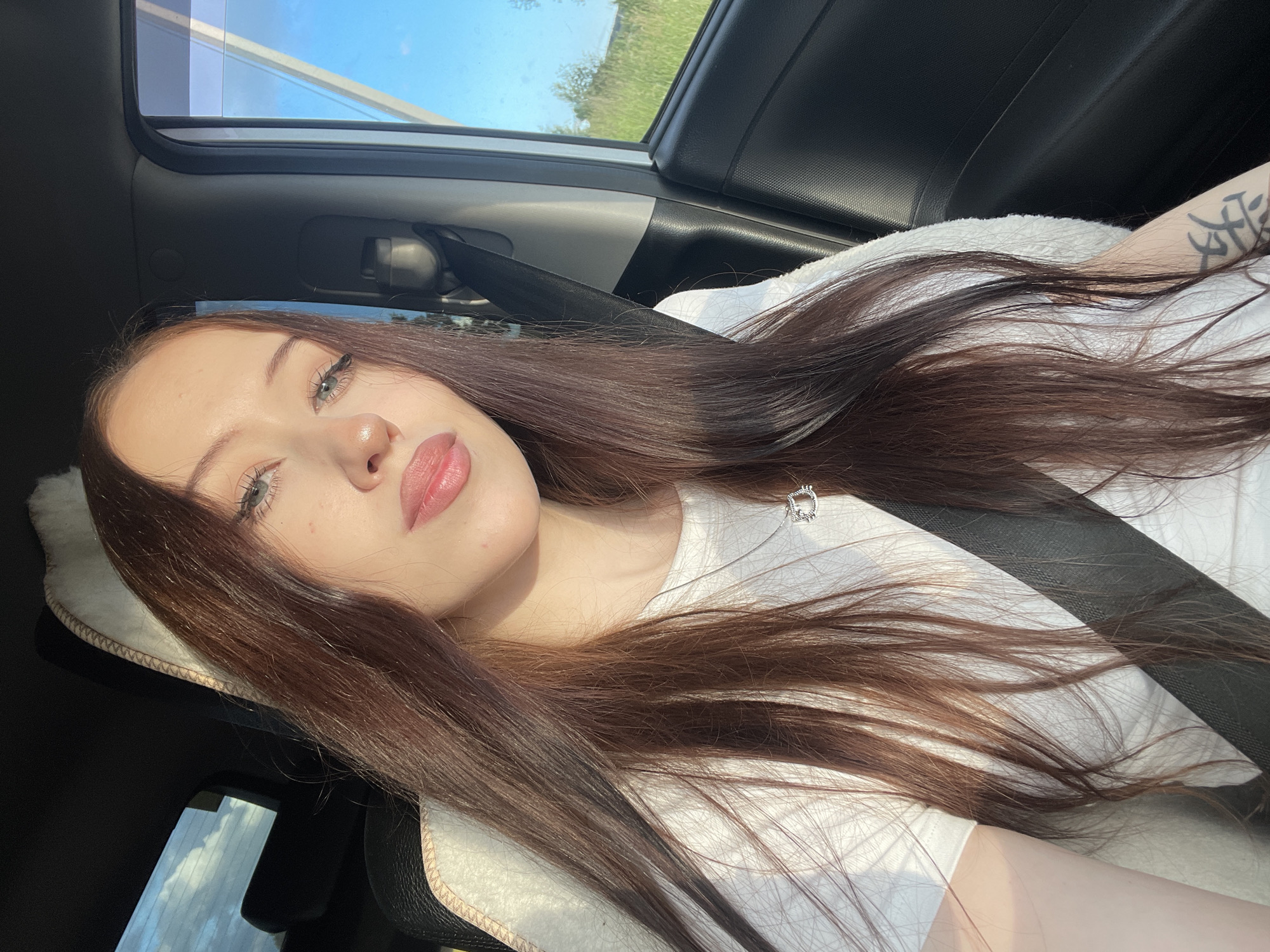 A young woman with long, dark brown hair is taking a selfie while sitting in the passenger seat of a car. The sun is shining brightly, casting shadows and highlighting her features. She is wearing a white t-shirt and a necklace with a small pendant. A seatbelt is visible across her chest. The interior of the car is dark, contrasting with the bright light coming through the car's sunroof, which shows a clear blue sky and green foliage outside. A tattoo is visible on her right arm. The overall mood appears relaxed and casual. The location is identified as Gavrilovka, Russia, and the time of day is likely midday due to the strong sunlight.