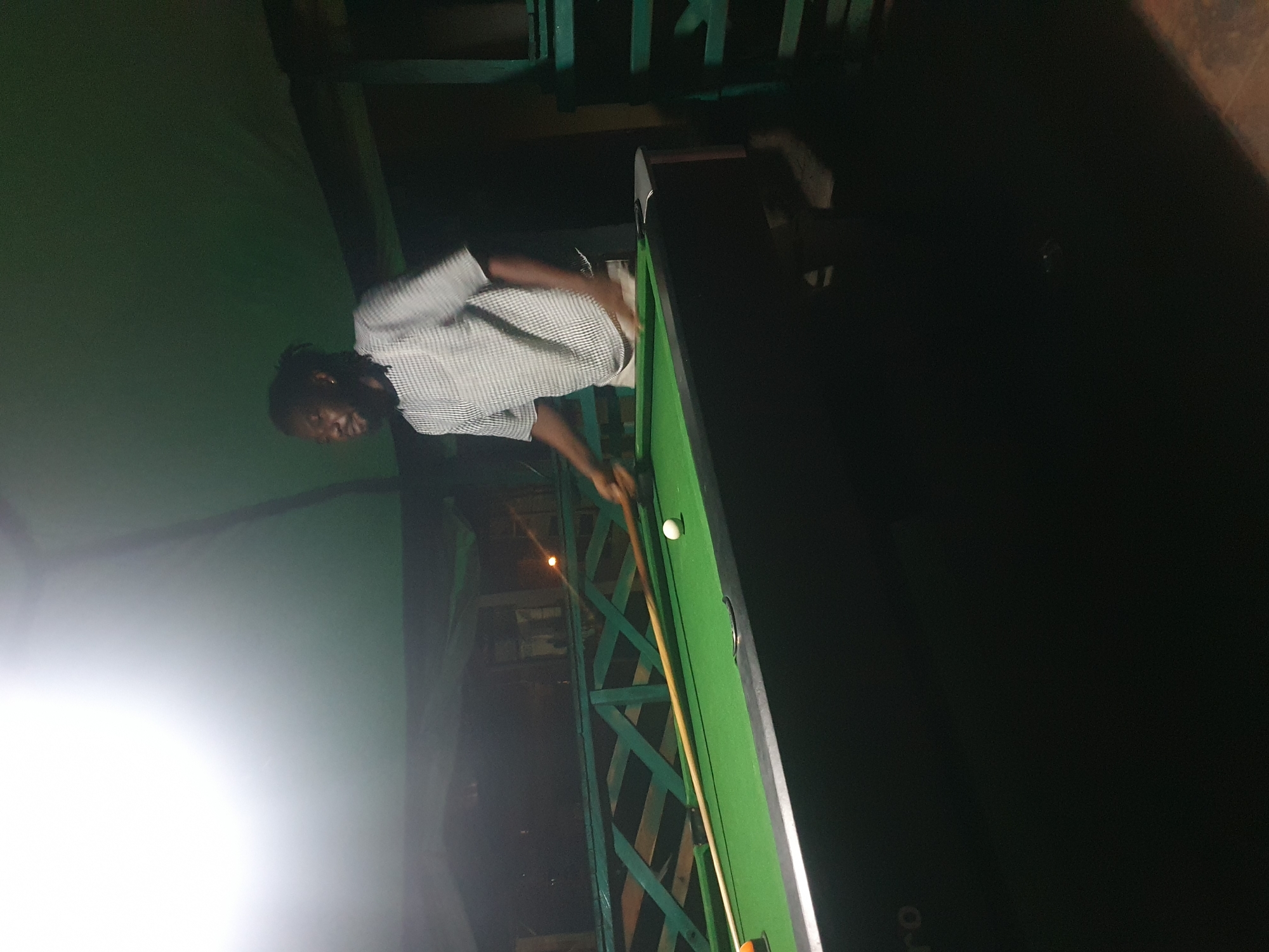 A man with dark skin and dreadlocks, wearing a checkered short-sleeved shirt and light-colored shorts, is playing pool. He is holding a pool cue and appears to be mid-shot. The pool table is covered in green felt, with a white cue ball and an orange object ball visible. The scene is outdoors at night, possibly at a bar or recreational area. A green wooden railing is visible in the background, and the lighting suggests it is under a roof structure. The atmosphere is casual and recreational. The provided hints confirm the subject is playing pool, and the environment appears to be Lokoja, Nigeria.