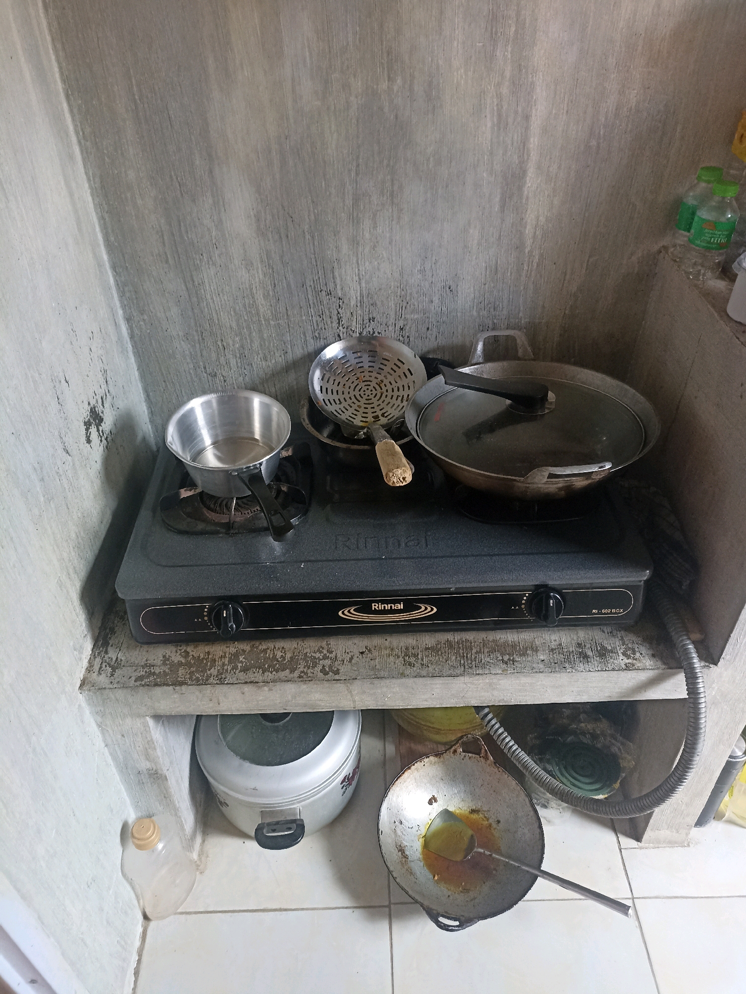 The image shows a corner of a kitchen with a concrete wall and tiled floor. On a concrete shelf, there is a black Rinnai brand gas stove with two burners. On the left burner, there is a small aluminum pot filled with liquid. On the right burner, there is a large, shallow pan with a glass lid. Next to the pot, there is a metal strainer with a wooden handle. Underneath the shelf, there is a silver rice cooker. To the left of the rice cooker, there is a clear plastic bottle with a yellow cap. In the lower right corner of the shelf, a metal spatula with a long handle rests in a dirty pan containing cooking oil. Two green-capped plastic bottles are visible on a higher ledge to the right, and a coiled mosquito repellent is partially visible under the shelf on the right. The overall scene depicts a functional but somewhat cluttered cooking area. The scene is located in the city of Mojopanggung, Indonesia.