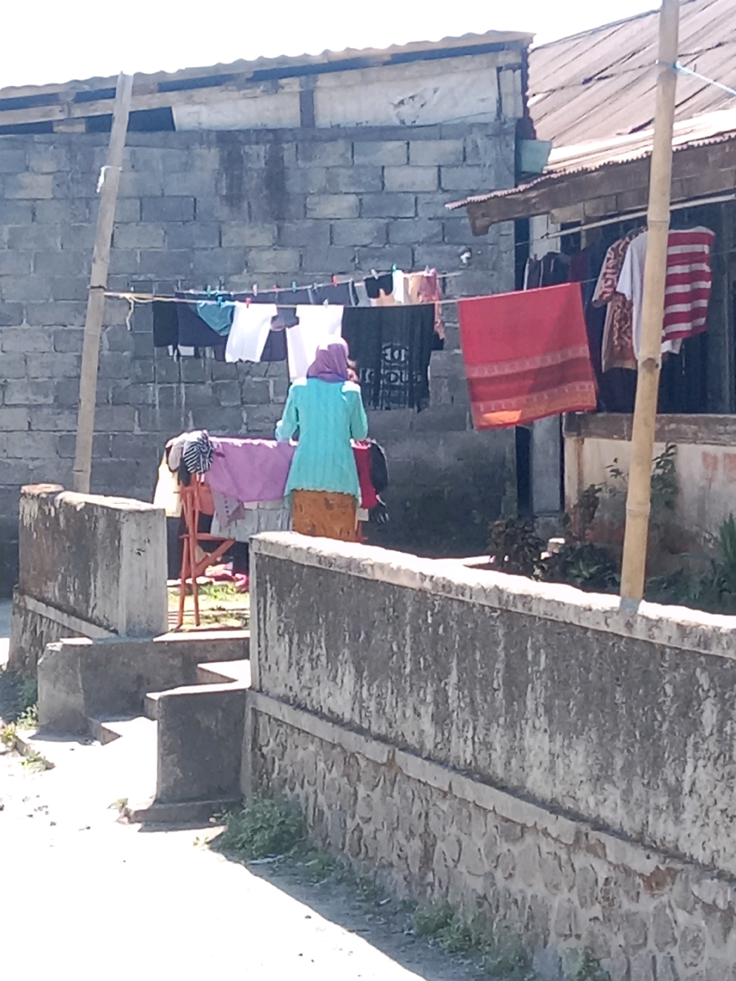 This image depicts an outdoor scene with a woman in the foreground, facing away from the camera, standing near a clothesline. The woman is wearing a blue tunic and a purple headscarf. The clothesline is strung between two poles and is laden with various garments, including shirts, pants, and a red cloth. Behind the clothesline is a grey brick wall, and to the right of that, a house with a corrugated metal roof. The ground appears to be dirt and gravel, with some patches of grass. The overall impression is one of a residential area, likely in a village or rural setting, given the rustic appearance of the structures and the outdoor drying of laundry. The presence of the brick wall and the simple house suggest a modest dwelling. The image quality is moderate, with some graininess and a slightly washed-out appearance, possibly due to bright sunlight. The location is specified as Jurangjeru, Indonesia.