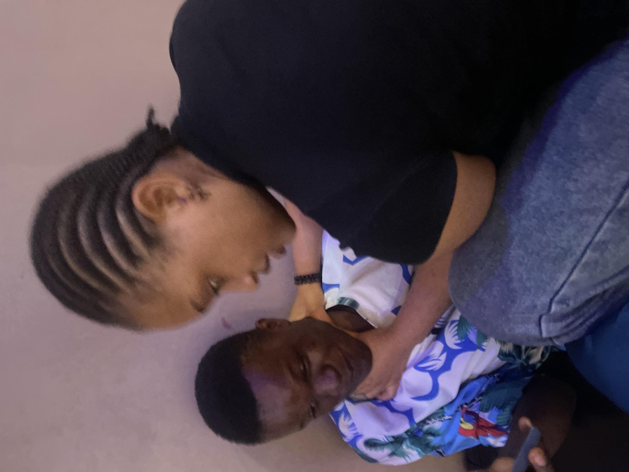 In Nkpor, Nigeria, a close-up shot captures two individuals. A young person with braided hair and a black shirt is leaning in, their hand gently touching the chin of another person. This second individual, wearing a white shirt with a blue and green floral pattern, is looking down, their expression indistinct. The background is a plain, muted wall. The image is shot from a slightly high angle, focusing on the interaction between the two people.