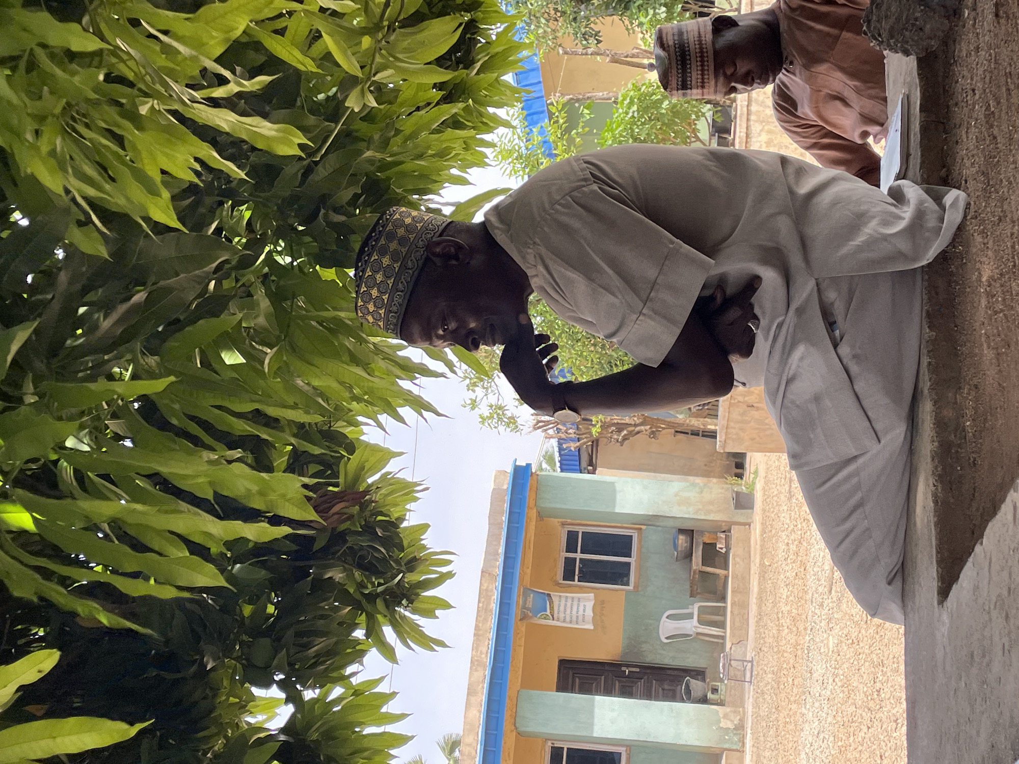 The image depicts an outdoor scene in what appears to be a residential or semi-urban area, consistent with Bukuru, Nigeria, under bright daylight with clear skies.
In the foreground, slightly to the left, a dark-skinned man is visible from the chest up. He wears a light-colored (possibly off-white or light grey) traditional long-sleeved garment and a patterned kufi cap, featuring geometric designs in gold, black, and light blue/grey. His head is bowed, and his hands are clasped or held together near his chin, suggesting a moment of prayer, contemplation, or deep thought. His expression is serious and introspective.
To the upper right, partially obscured, another dark-skinned man is visible, wearing a light brown or beige traditional garment and a striped cap. He appears to be looking down at something, possibly a surface in front of him, and seems engaged in an activity.
Lush, green foliage, likely from a tree, dominates the left side and upper portion of the image, suggesting a natural, shaded environment. In the background, multi-story buildings are visible. One prominent building features a light yellow/orange lower section and a light blue/green upper section, with white-framed windows and a dark brown door. A bright blue horizontal stripe or trim runs across the building's facade. A white plastic chair is visible near the buildings.
On the building in the background, a white sack or piece of fabric hangs, displaying black text. Visible words include "TAL PEPPER MIX", "RED PEPPER MIX", "DRY GINGER", and "BEEF MEAT", indicating a list of food products or ingredients.