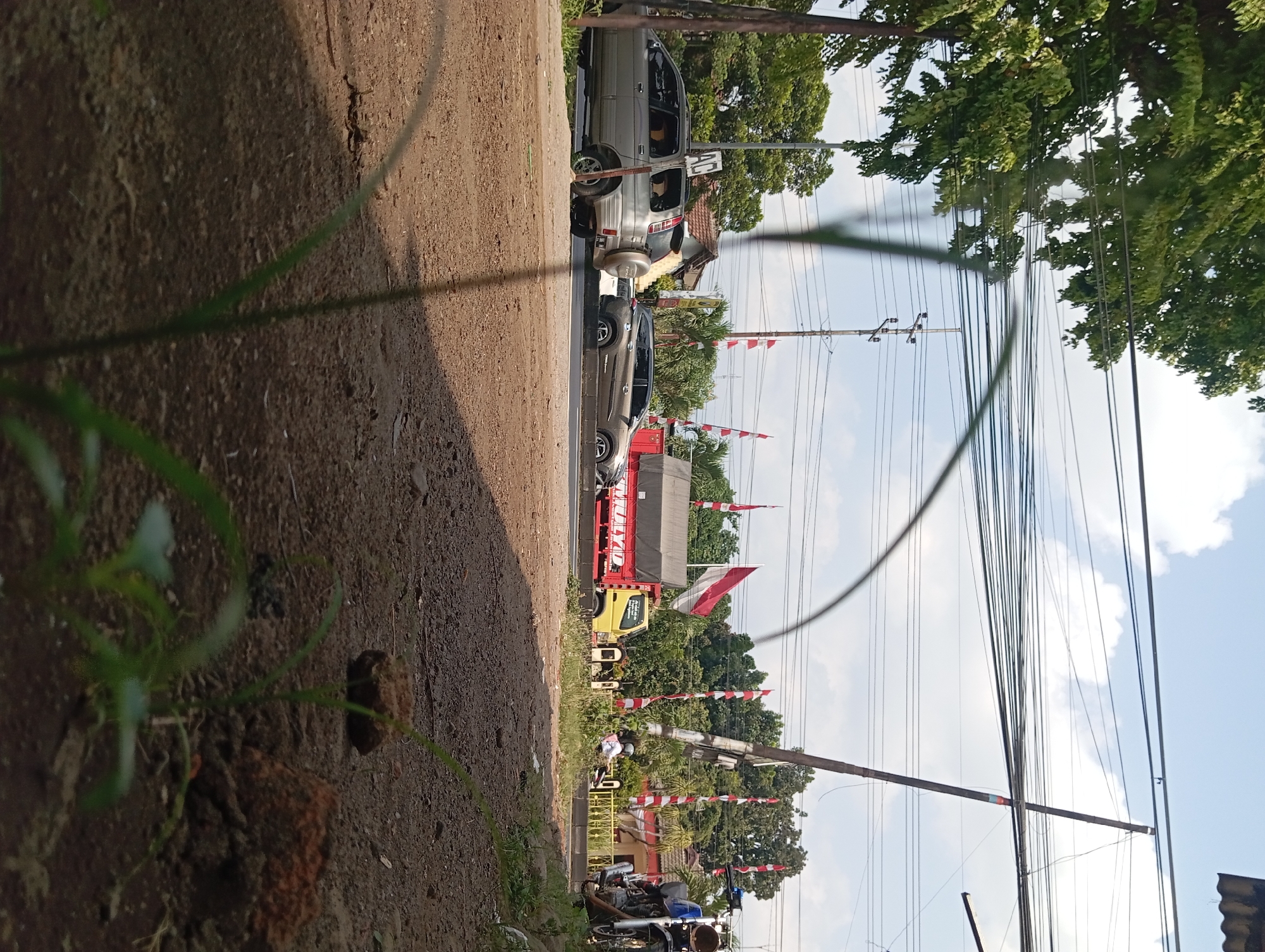 The image depicts a street scene in Lemahbang, Indonesia, on a sunny day with scattered clouds. In the foreground, the ground is composed of dry dirt and small rocks, with blades of grass growing sporadically. A low-angle perspective looking towards the street is used.
On the street, several vehicles are visible. A silver SUV is parked on the left side of the road, facing right. To its right, another car, a dark brown SUV, is driving on the road. Further down the street, a yellow truck with a red cargo bed is stopped. A motorcycle is parked on the right side of the frame, partially obscuring the view of the street.
The environment includes trees and vegetation along the side of the road. There are also power lines and utility poles overhead, indicating an urban or semi-urban setting. Red and white flags, commonly used for national celebrations in Indonesia, are displayed on poles along the roadside, suggesting a festive or commemorative occasion. A few people are present, including a person on a motorcycle and others in the background near buildings. The overall impression is a busy street with everyday activity, overlaid with elements of local decoration.