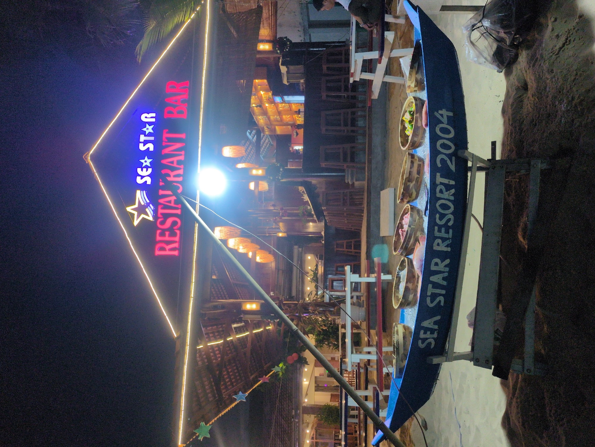 The image depicts an outdoor scene at night, likely at a tourist destination. A prominent feature is a sign for "SEA STAR RESTAURANT BAR," illuminated with neon lights in red, blue, and yellow. The sign is shaped like a triangle and is the focal point of the image.
In the background, there is a building or structure with a thatched roof, possibly a restaurant or bar area. Hanging lanterns provide ambient lighting. The presence of tables and chairs suggests a dining area.
To the right of the frame, there is a blue boat labeled "SEA STAR RESORT 2004". There are woven baskets placed inside the boat.
The ground appears to be sandy, indicating a coastal or beach location.
The location is likely in Phú Quốc, Vietnam, based on the name of the establishment.