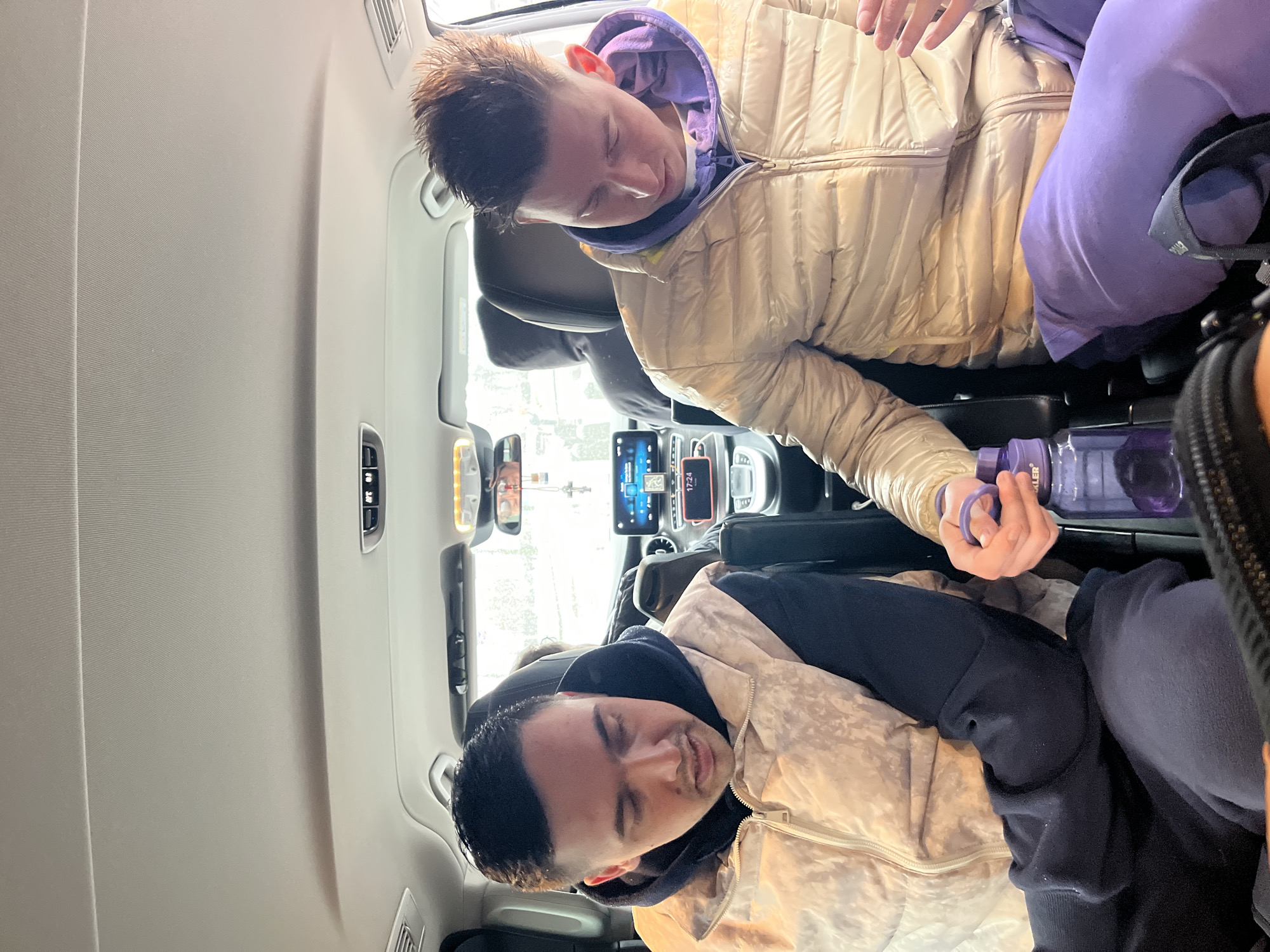 Two males are seated in the back of a vehicle. The male in the upper portion of the frame wears a beige puffer jacket over a purple hooded sweatshirt and purple sweatpants. He appears to be holding a purple water bottle. The male in the lower portion of the frame wears a light brown and beige camo print jacket over a dark hooded sweatshirt and dark sweatpants. The vehicle's interior includes a rearview mirror, dashboard screen, and climate control panel. Based on the interior design and features visible, the scene is likely inside a modern passenger vehicle, but no information can be gleaned about the exact location or whether it is in Moscow, Russia.