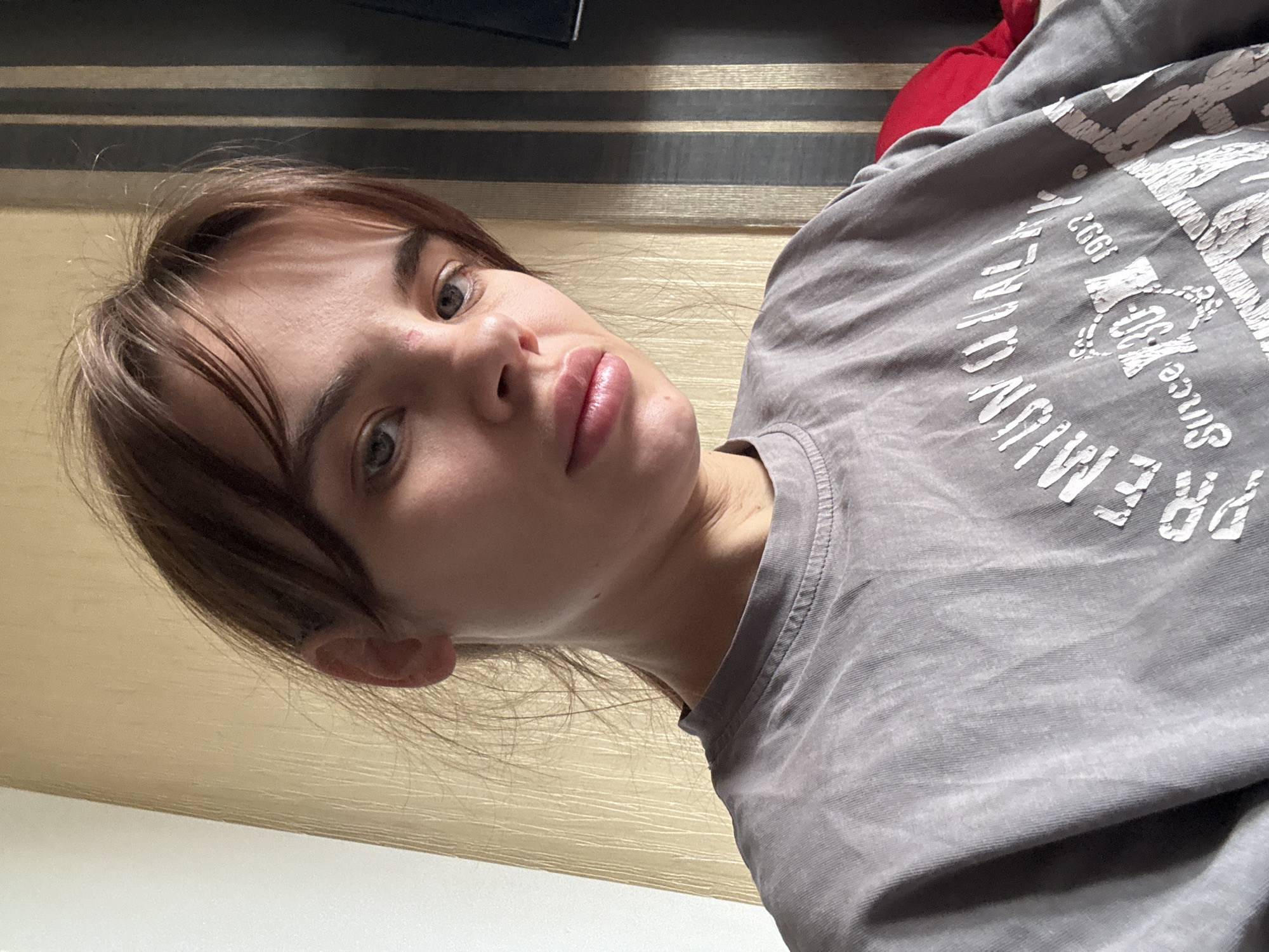 A young woman with fair skin and brown hair with bangs is pictured in a selfie-style image. She is wearing a gray long-sleeved shirt with white text. The background includes a striped wall and a partially visible red object. The location is possibly indoors, but the specific city, Rostov-on-Don, Russia, cannot be confirmed from the image alone. No specific actions or interactions can be determined.