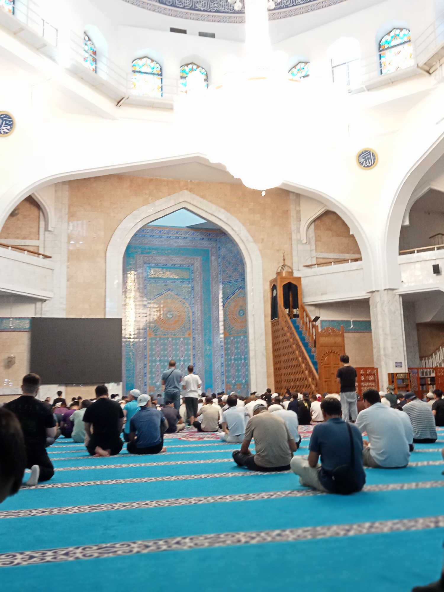 The image depicts the interior of the Central State Mosque in Almaty, Kazakhstan, during a prayer service. A large congregation of men are seated on a blue carpet, which is divided by white stripes. The mosque's architecture is grand, with white arched ceilings and walls decorated with intricate blue tilework. A tall wooden minbar, a pulpit for sermons, is visible on the right side of the image. A large screen is positioned on the left side of the mosque. The lighting is bright, suggesting it is daytime. The men are dressed in casual clothing and appear to be engaged in prayer, with many facing towards the qibla (direction of prayer). The overall atmosphere is one of solemnity and devotion.