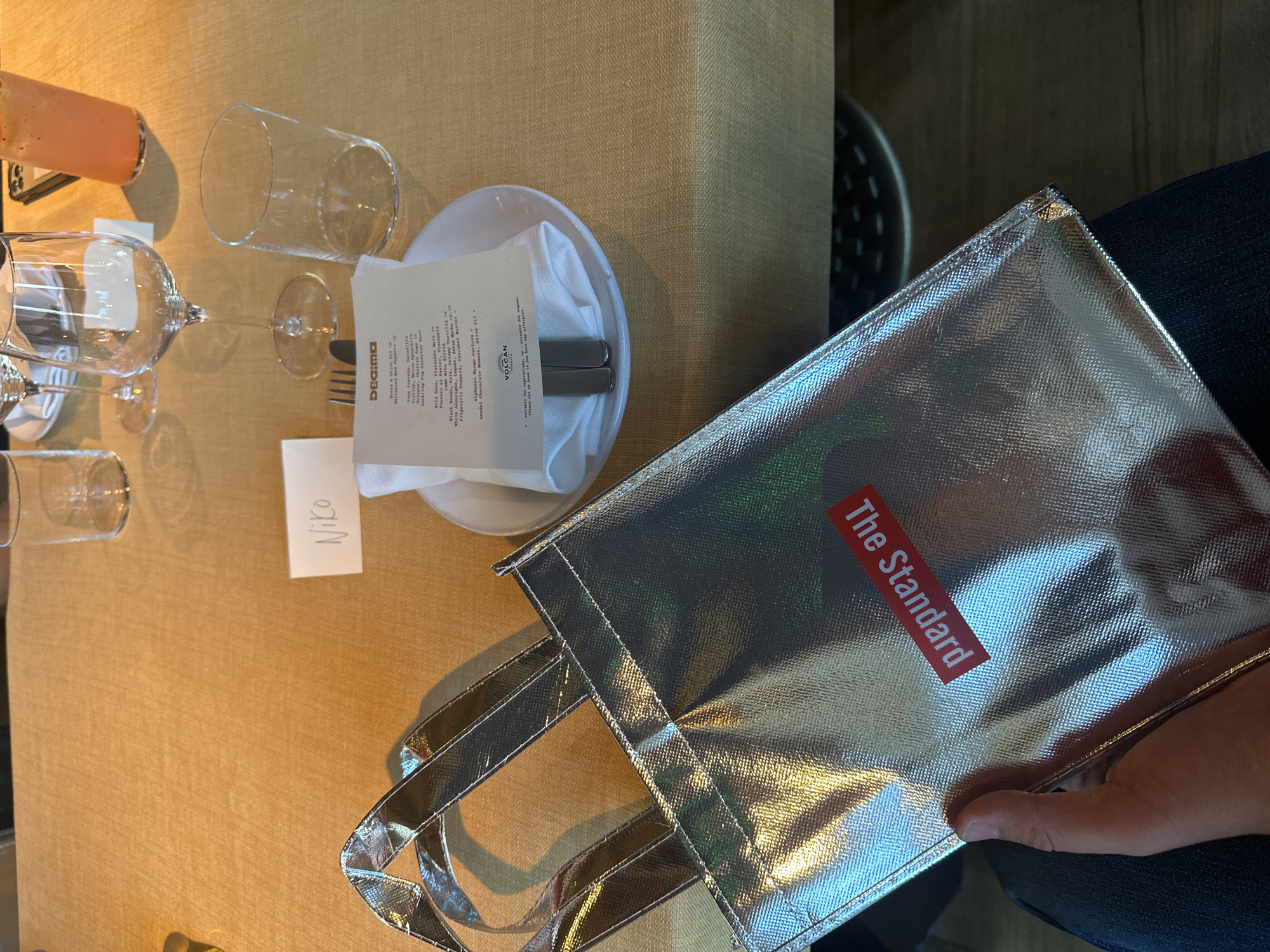 The image shows a table setting, likely in a restaurant named "Decimo Volcan" (London, UK) based on the menu. The table is covered in a tan tablecloth and includes wine glasses, a water glass, a juice glass, a plate with silverware and a napkin, and a place card with "Niko" written on it. A shiny silver bag with "The Standard" printed on it in red is also on the table, partially held by a hand. The bag suggests a possible connection to The Standard hotel, which has a restaurant named Decimo.