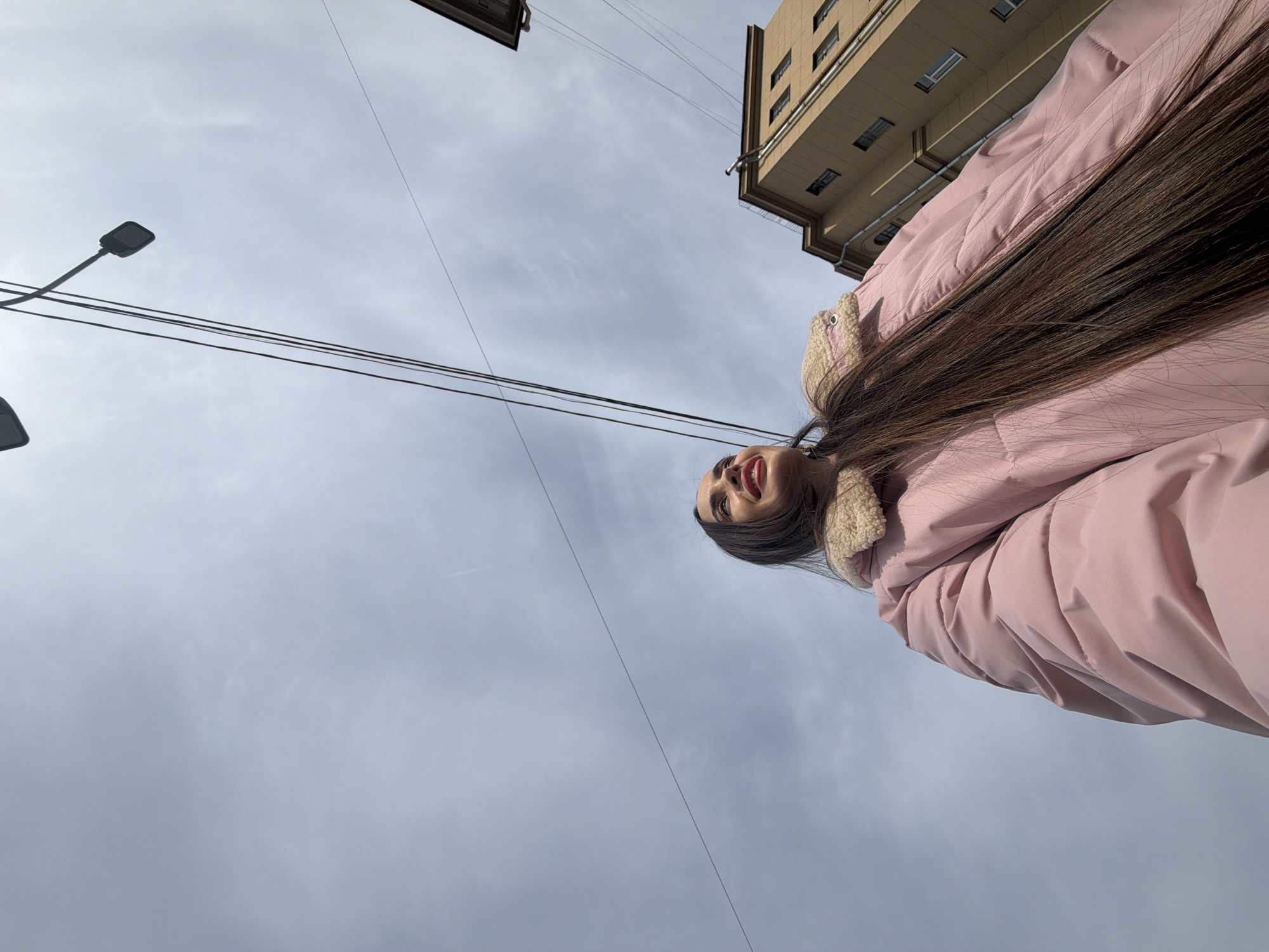 A low-angle shot of a young woman with long brown hair, wearing a pink puffer jacket with a beige sherpa collar, against a cloudy sky. She is looking upward, smiling, with a portion of a beige building visible in the upper right and a black street lamp on the upper left. Electrical wires cross the frame diagonally. The photograph was taken in Saint Petersburg, Russia.