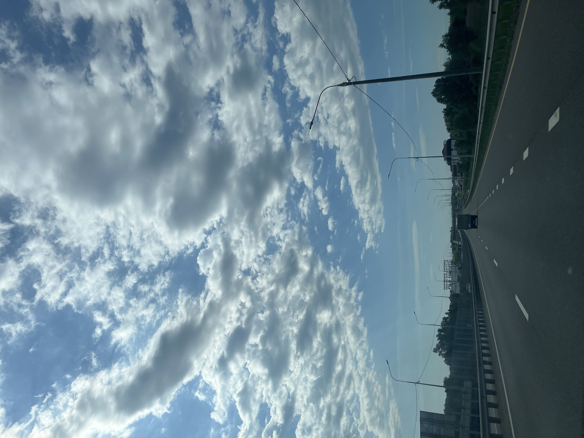 The image shows a highway in Psekups, Russia. The road is gray asphalt with white lane markings. On the right, there is a metal guardrail, followed by a strip of green vegetation and scattered buildings. Along the left side of the road, lampposts stretch into the distance. A dark car is visible further down the highway. Above, the sky is blue, partially covered by scattered white clouds.