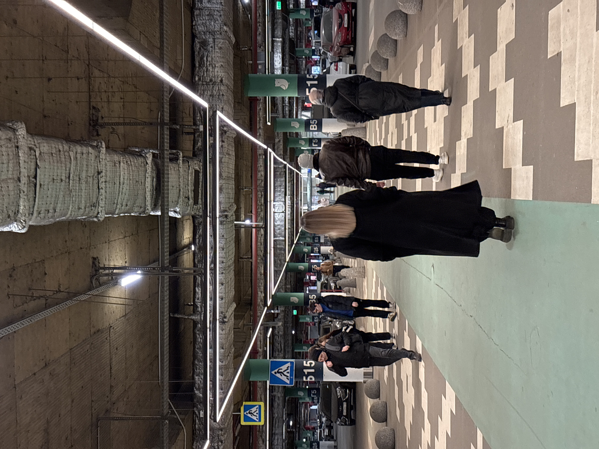 The image shows an underground parking garage in Kudrovo, Russia. Several people are walking along a pedestrian walkway marked with a green stripe. The walkway is adjacent to parked cars. Columns are spaced along the walkway, with signage indicating parking space numbers (e.g., "Б515"). The floor is patterned with alternating light and dark grey stripes. Overhead, there are bright, linear LED lights in a rectangular pattern, and insulated pipes are visible on the garage ceiling and walls.