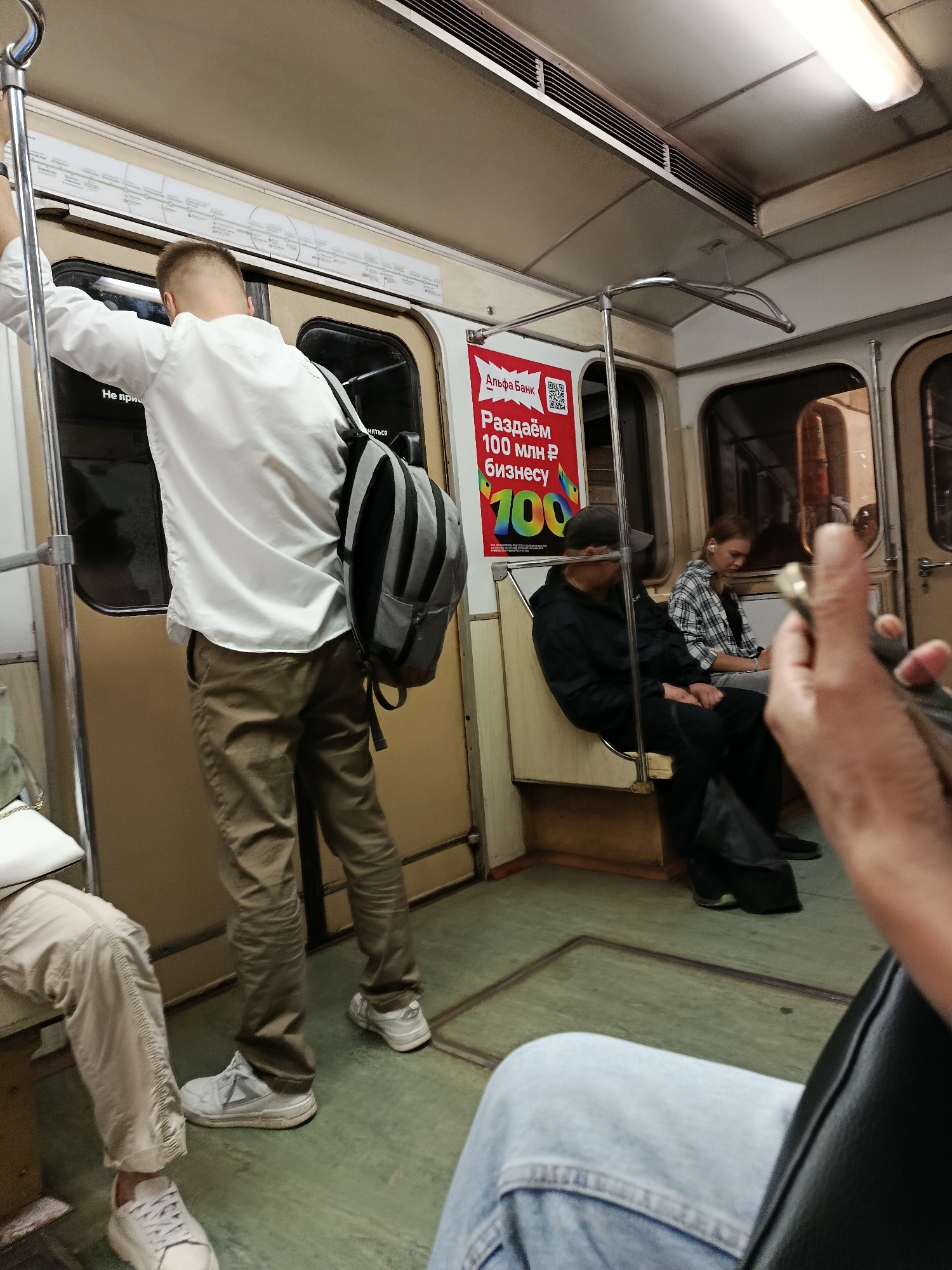 In the interior of a subway car, a young man in a white shirt and khaki pants stands, holding a pole. He wears a gray and black backpack. Several other passengers are seated, including a man in a black hoodie and baseball cap, a woman in a plaid shirt, and a person in light-colored pants on the left. Another person's hand, holding a phone, is visible in the foreground on the right. The car is beige and brown, with green flooring. A red advertisement with Russian text and a QR code is posted on the wall.