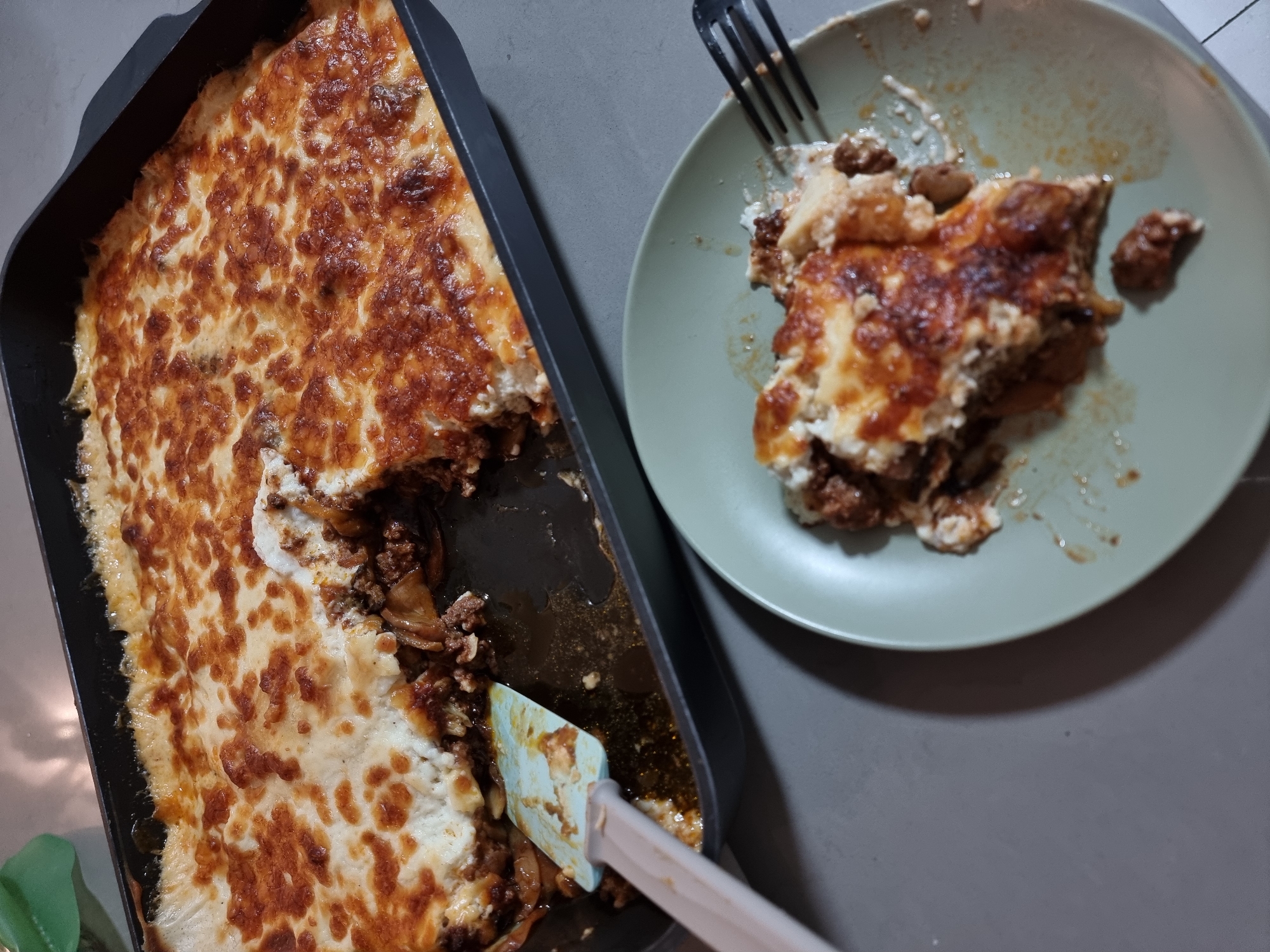 The image displays an overhead view of a hearty casserole dish, partially served, resting on a plain grey surface, likely a kitchen counter or table. On the left, a large, black rectangular baking pan holds the bulk of the casserole. The dish features a golden-brown, bubbly, and slightly crisped top layer, suggesting melted cheese or béchamel sauce. Below the browned crust, visible where a portion has been removed, are layers of dark ground meat, possibly interspersed with vegetables. A light blue and beige spatula is inserted into the casserole within the baking pan, indicating an ongoing serving process.
To the right, a single, light green or sage-colored round plate contains a serving of the same casserole. A black fork, with remnants of food, rests on the plate, which also shows streaks of sauce and grease from the meal. The food on the plate appears warm and ready for consumption.
The lighting is consistent with an indoor setting, likely overhead. No people are visible in the frame, nor is there any discernible text. The scene evokes a comfortable, domestic mealtime ambiance. The location context is Ramat Gan, Israel.
