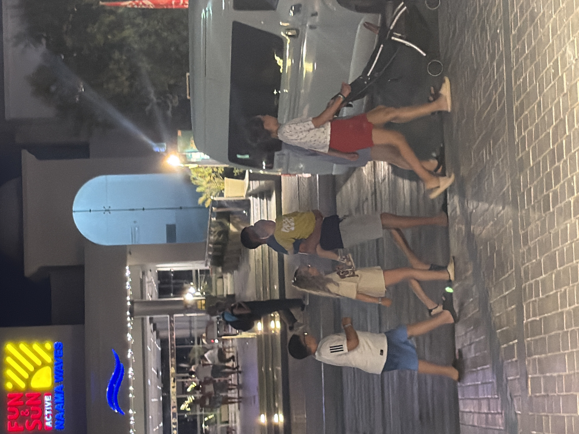 A group of people are walking along a paved walkway at night, likely in Sharm El Sheikh, Egypt, near the "FUN & SUN ACTIVE NAAMA WAVES" resort. In the foreground, a young boy wearing glasses and a white t-shirt with black stripes, along with blue shorts, walks to the left. Beside him, a young girl with blonde hair, wearing a beige t-shirt and shorts, walks with him. A man in a blue shirt is carrying a toddler in a yellow shirt and grey shorts. To the right, a girl in a white patterned t-shirt and red shorts is pushing a stroller. Behind them, a white van is parked. In the background, lights illuminate the entrance of a building with an arched entryway, and a blue wave-like sculpture is visible above. Other individuals and an illuminated walkway can be seen in the distance. The scene suggests a casual evening stroll.