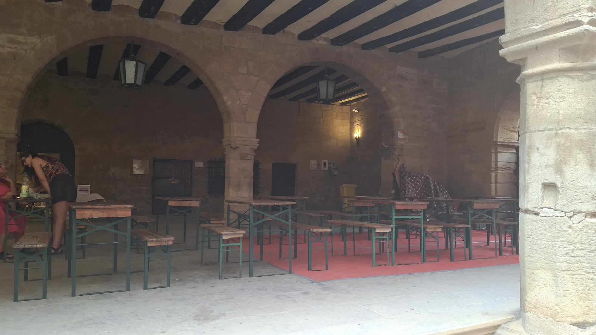 In La Fresneda, Spain, an outdoor area with stone arches and wooden beamed ceilings features several picnic tables with benches set up on a red carpet. A woman wearing a patterned top and black skirt is standing at one of the tables, leaning over it. Another person, wearing a red garment, is seated at a table to the left, partially visible. The setting appears to be an establishment or event space, possibly a bar or restaurant, given the presence of tables and seating arranged for patrons. The architecture suggests an older, historical building.