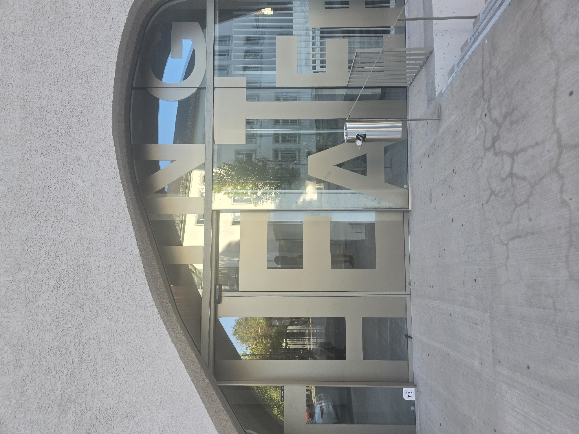 The image shows the facade of a building with large, metallic letters spelling out "THEATER" across the glass entrance. The letters are silver and appear to be part of the building's design. The building has a modern architectural style with a textured, off-white exterior wall above the glass. Reflected in the glass are trees, buildings, and the sky. To the right of the entrance, a metal railing leads down a set of stairs. A cylindrical metal trash receptacle is positioned near the entrance. The ground in front of the building is paved with concrete. The setting appears to be a public space in Saarlouis, Germany, given the context.