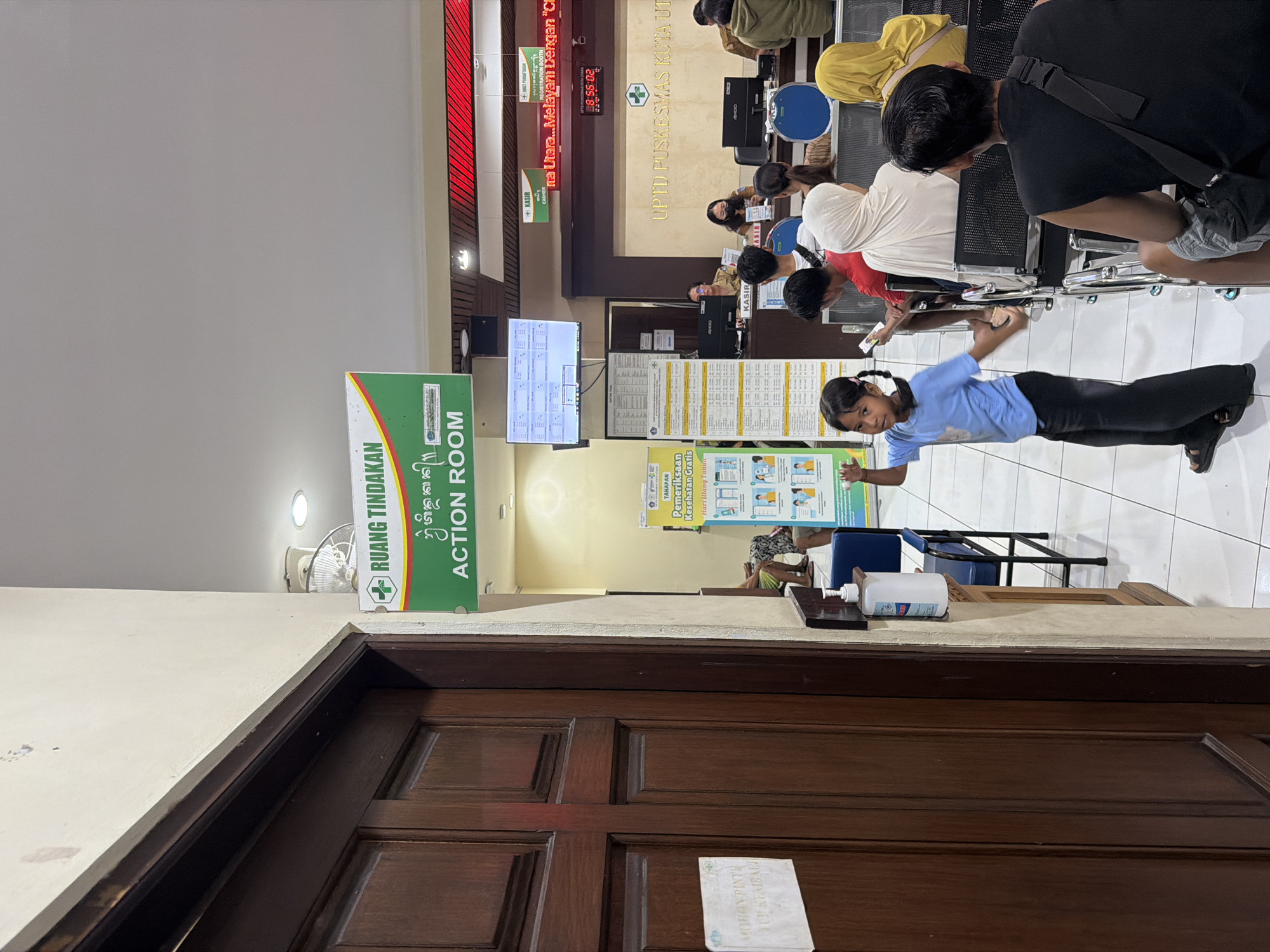 The image captures the interior of a healthcare facility, specifically the UPTD Puskesmas Kuta Utara in North Kuta, Indonesia. A young girl stands in the foreground, facing the camera, wearing a blue shirt and dark pants. Behind her, patients are seated in rows, waiting for service. At the service counter, healthcare workers attend to the patients. Prominently visible is a sign indicating the "RUANG TINDAKAN ACTION ROOM." Additional signage displays information about free health check-ups and other health information. Digital clocks displaying time. A door with signage is visible in the foreground with a bottle of sanitizer. Monitors display patient queuing information.