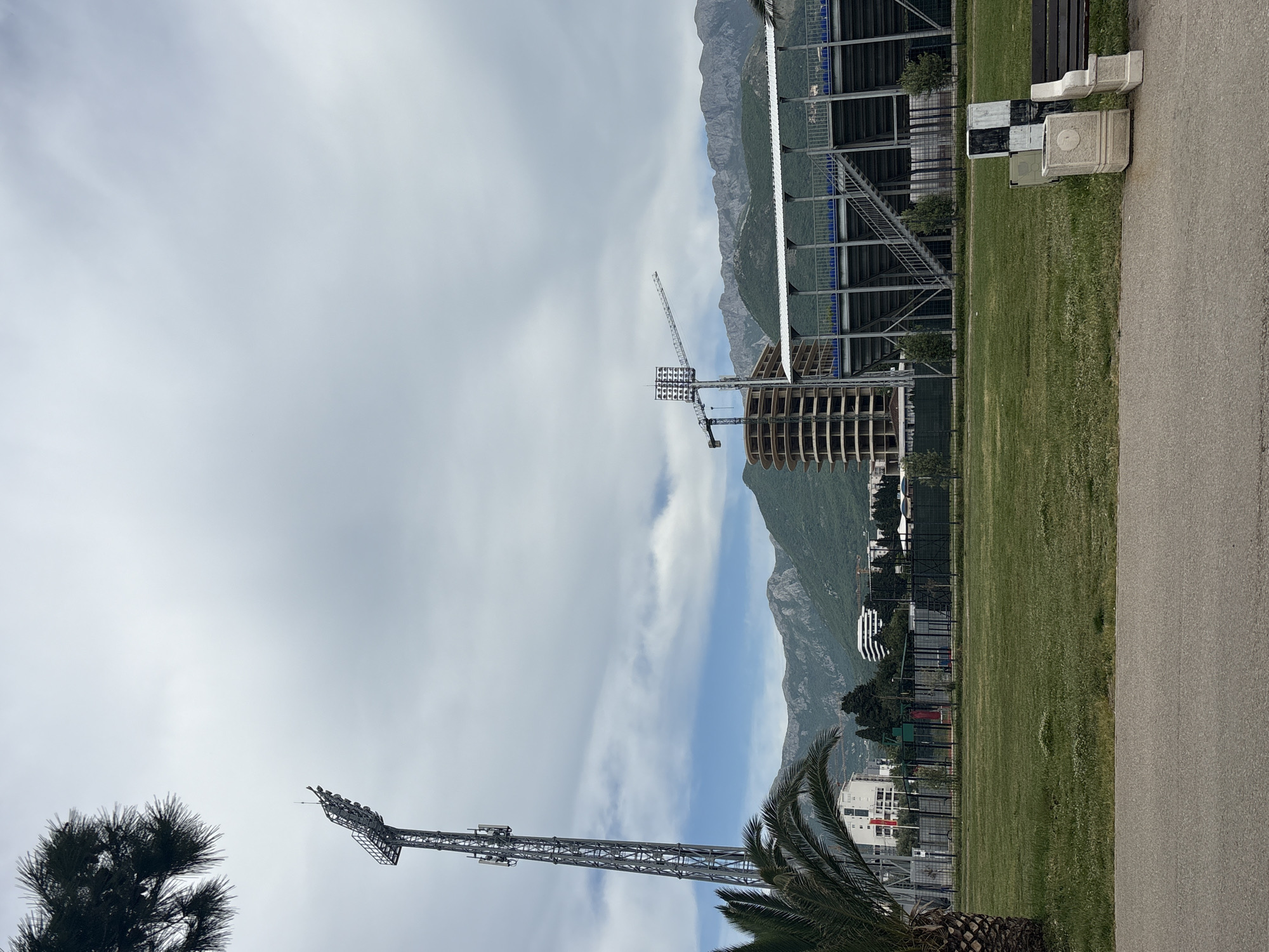 The image depicts a construction site in Bar, Montenegro, set against a backdrop of mountains and a cloudy sky. A partially constructed building stands in the center of the frame, accompanied by a construction crane. A tall floodlight tower is visible in the foreground, near a palm tree. Adjacent to the construction site is a sports stadium, suggested by the presence of stadium seating and fencing. A paved walkway runs along the right edge of the image, with benches and possible construction materials nearby. The overall environment suggests an urban setting undergoing development near natural terrain.