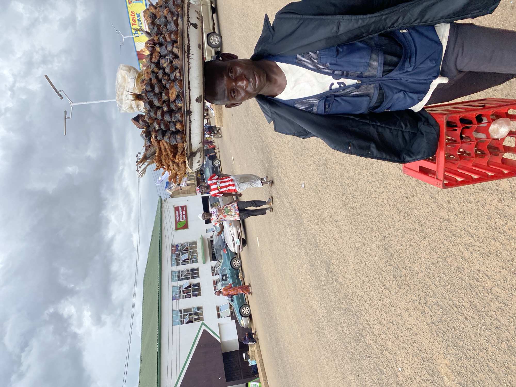 The image shows a street scene in Bukuru, Nigeria, under a cloudy sky, likely in the daytime. A man is standing next to a truck filled with dried fish. He is wearing a dark jacket over a white shirt and a blue vest. He is looking towards the camera with a neutral expression. In front of him, a person in a red and white striped shirt and light-colored pants stands looking away from the camera. Behind them, a building with multiple windows and a green roof is visible, with a sign that reads "HIGHLAND MICROFINANCE BANK PLC." A red plastic crate filled with what appears to be bottles is placed on the ground near the man. Several cars and other people are also present in the background, suggesting a busy market or commercial area. The overall impression is of a typical street vendor scene in a Nigerian town.