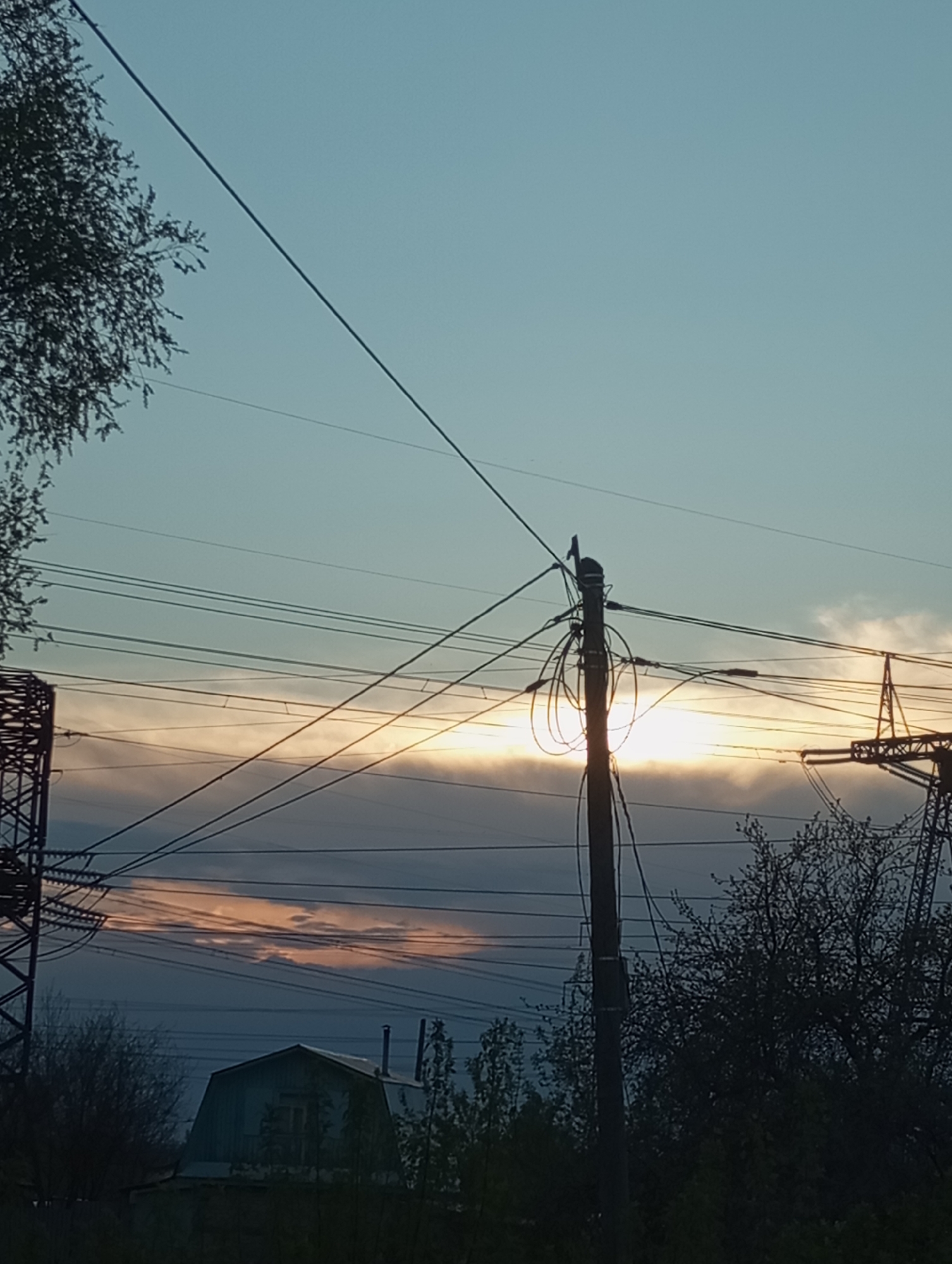 The image depicts a late evening or early morning scene in Chaikovsky, Russia. A low-rise building with a gable roof is visible in the foreground, obscured by trees and foliage. Power lines and electrical poles dominate the midground, silhouetted against a sky with hues of orange and blue visible through the clouds. The light source appears to be behind the clouds, possibly the setting or rising sun. The scene suggests an industrial or residential area on the outskirts of the city. There are no people visible in the image.