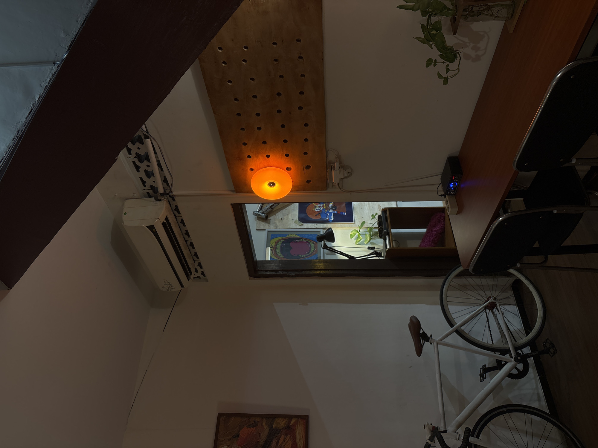 A dimly lit indoor scene, likely a residential or commercial space in Semarang, Indonesia, captures a corner of a room with a functional, somewhat eclectic arrangement of objects. A white bicycle is parked against a white wall on the left, its seat a brown leather. To the right of the bicycle, an open doorway reveals a glimpse into another room, where a black desk lamp illuminates a shelf with a plant and various items. Above the doorway, framed art is visible.
Dominating the right side of the image is a large, light-brown wooden panel mounted on the wall, featuring a grid of evenly spaced holes. A single, round, orange light fixture is attached to the panel, casting a warm glow. Below the panel, a dark brown rectangular table is set with two black chairs. On the table, a small black electronic device with blue indicator lights is placed next to a potted plant with green vines in a glass vase. The ceiling shows an air conditioning unit and a dark wooden beam. The overall impression is one of a lived-in space, potentially a cafe, studio, or home, characterized by a mix of modern and vintage elements.