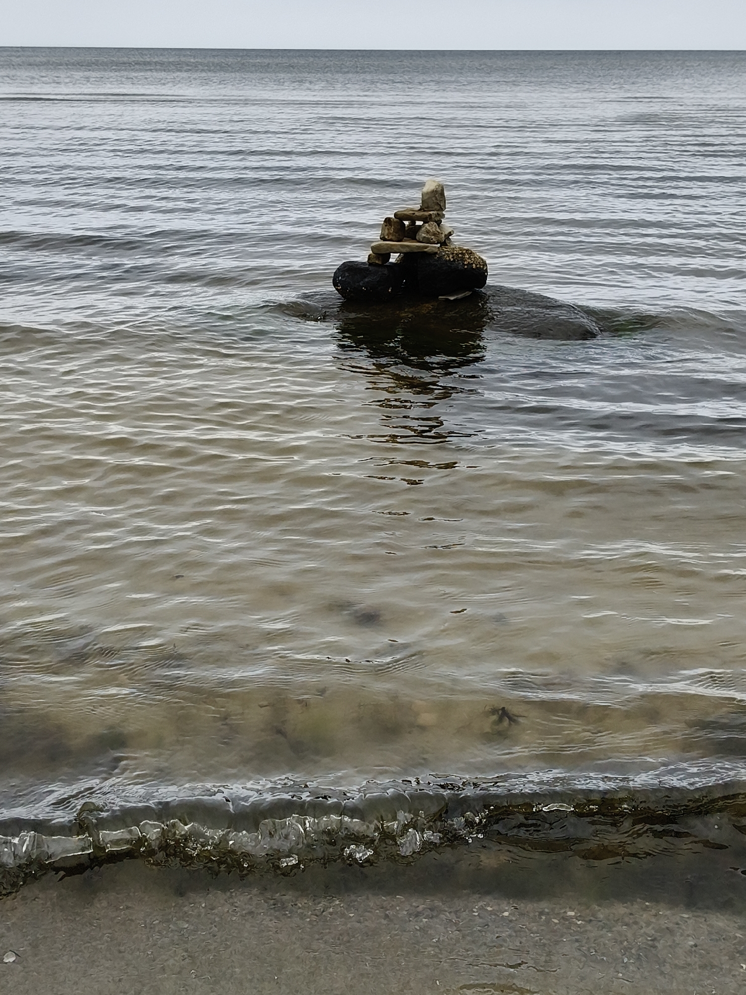 An inukshuk made of stacked stones stands on a dark, partially submerged rock in the calm, shallow waters of the Baltic Sea at Bigauņciems, Latvia. The water is clear enough to reveal the sandy seabed and scattered pebbles beneath the surface. Small waves gently lap at the shore in the foreground, where a pebbly beach meets the water. The sky above is overcast and gray, with the horizon line clearly visible where the water meets the sky. The scene is devoid of any people or other living creatures.