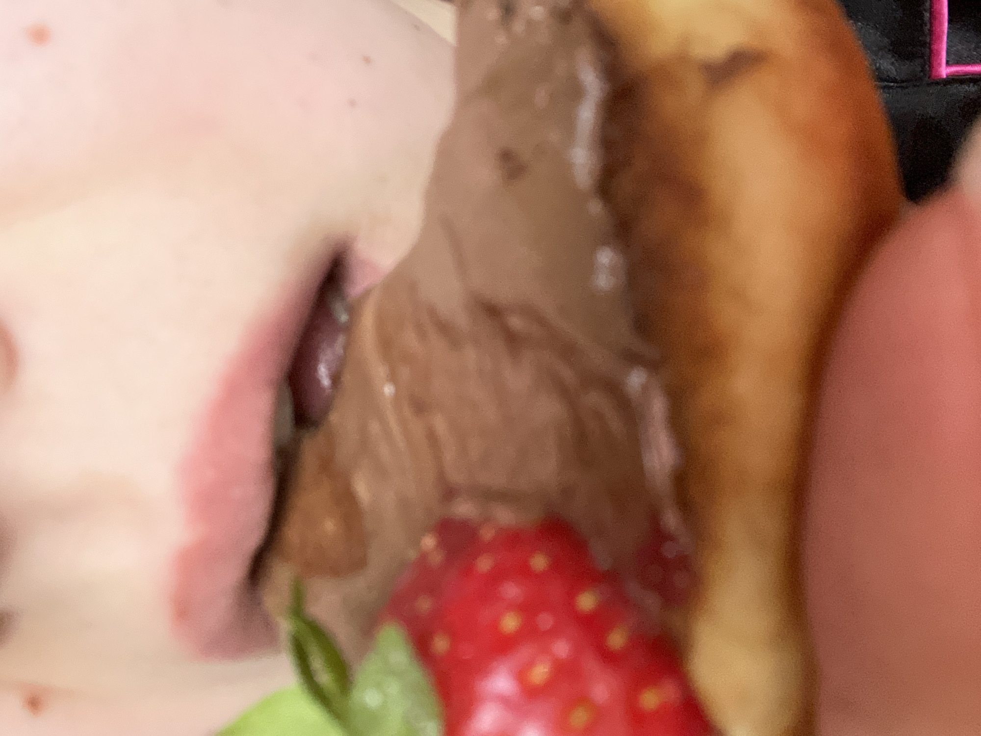 This close-up image captures a person eating a dessert. The left side of the frame features the lower portion of a person's face, including their chin and open mouth, revealing a pink tongue. The skin is fair, with a few visible freckles or moles. A generous amount of rich, brown, creamy substance, likely chocolate ice cream or a similar dessert, is being consumed, with some of it smeared around the mouth.
To the right, partially covered by the chocolate, is a golden-brown baked item, resembling a donut or pastry. At the bottom center of the frame, a vibrant red strawberry with its distinct seeds and green leaves is clearly visible. The image has a soft focus, emphasizing the textures of the food and the intimate act of eating. In the top right corner, a blurry dark object with a faint pink stitch or embroidery is present, but no discernible text can be made out. The scene depicts a moment of enjoying a sweet treat. While no explicit location cues are visible within the image, the context provided indicates the location is Plovdiv, Bulgaria.
