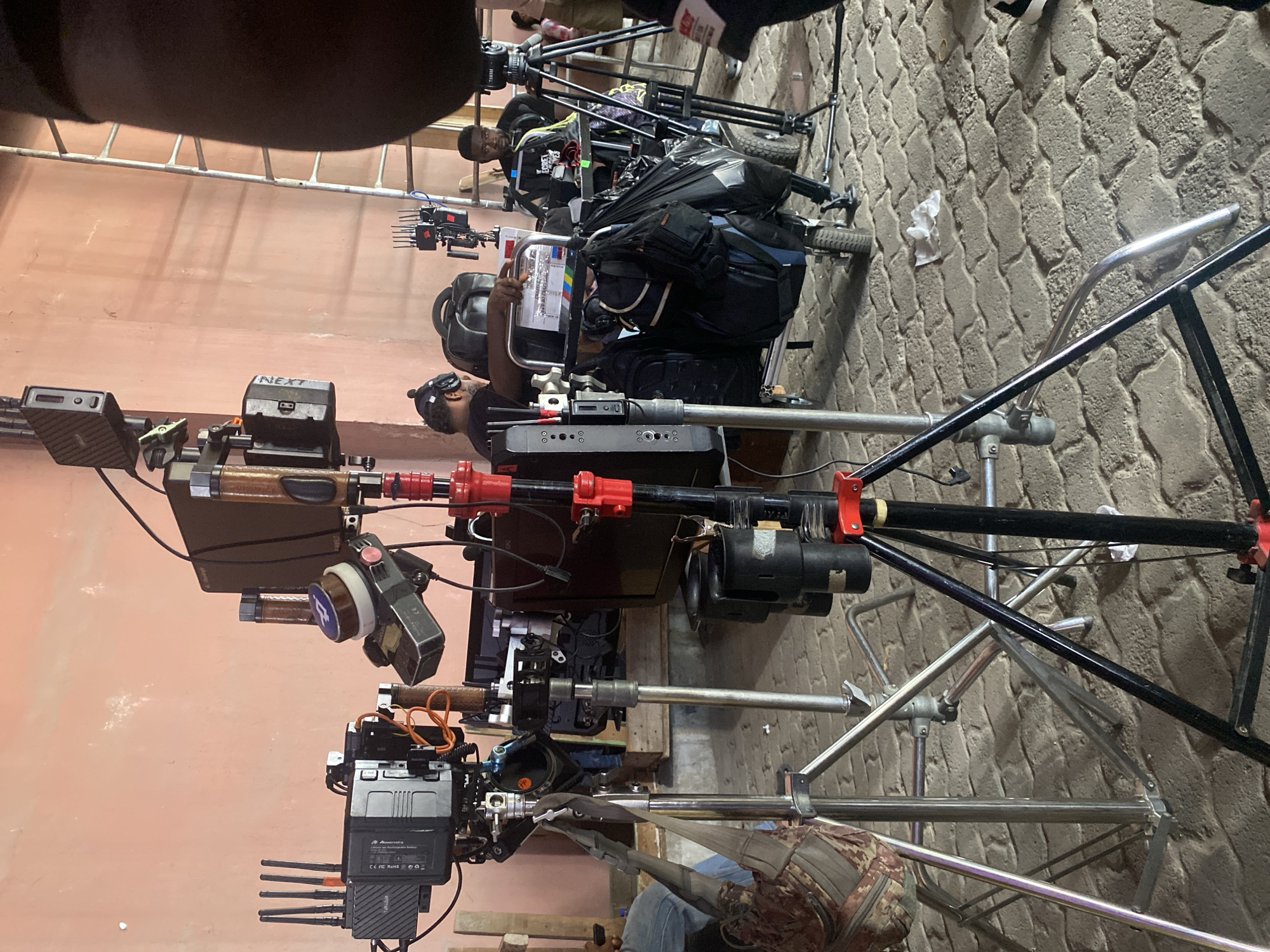 The scene shows a film set in Lagos, Nigeria. Multiple camera rigs are visible, each mounted on tripods. Two people are present: one holding a clapperboard, the other operating a camera. The rigs are equipped with various accessories, including monitors, wireless video transmitters, and lens control systems. The ground is paved with patterned stone. The background shows a plain painted wall.