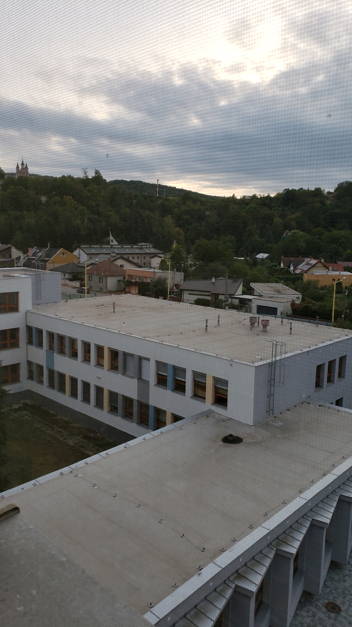 An aerial view captures a gray, cloudy sky over a residential and institutional area. The foreground features a flat, light-colored roof with visible metal seams and fasteners, part of a modern building. Beyond this roof, a larger, multi-story building with a light gray facade and numerous windows is visible. These windows have orange, blue, and yellow frames. A metal ladder is attached to the side of this building.
In the middle ground, a cluster of houses with various colored roofs and facades, typical of a European town, are nestled amongst lush green trees. A prominent church with twin spires is situated on a hill in the distance, to the left of the frame, under the overcast sky. A telecommunications tower is also visible on another hill. The overall impression is of a quiet, somewhat overcast day in a European town. The image is consistent with a view from a building in the city of Prešov, Slovakia, possibly from a higher floor looking out over the surrounding landscape and neighboring structures.
