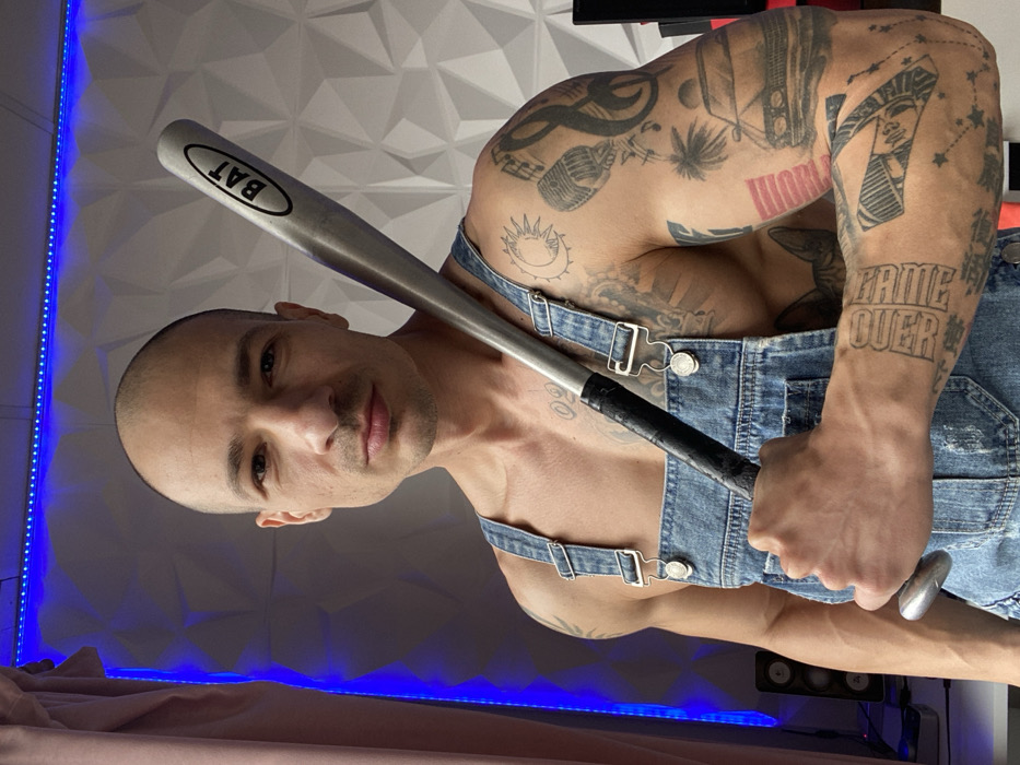 An adult male with a bald head, light skin, and a neutral expression stands indoors, looking directly at the camera. He is shirtless, wearing blue denim overalls with the straps visible, and exhibits a muscular build. Both arms are extensively covered in tattoos; the right arm (viewer's left) displays designs including a sun and moon, a microphone, musical notes, a car, and text, while the left arm (viewer's right) also features numerous tattoos, including text on the forearm. He holds a silver-colored metallic baseball bat with a black taped grip, resting it on his left shoulder. The bat's barrel clearly shows the word "BAT" enclosed within an oval. The background consists of a white, geometric-patterned wall, illuminated by blue LED strip lighting along its top and left edges. A light pink curtain is partially visible on the far left, and a dark electronic device is seen on the far right. A black speaker is positioned to the left of the male's waist. The scene is located in the city of Pleven, Bulgaria.