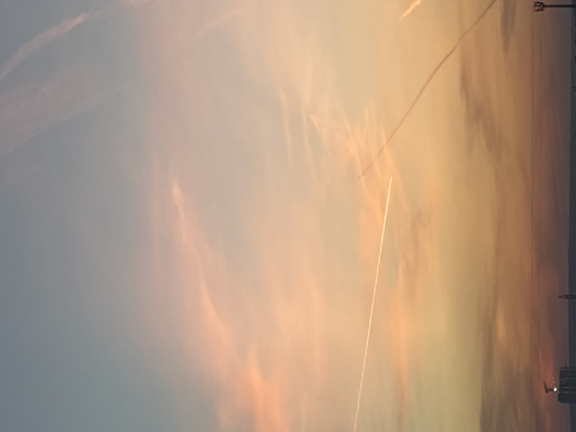 The image depicts a sky at sunset or sunrise, with hues of orange, pink, and blue blending together. Streaks or contrails are visible across the sky, likely from aircraft. The horizon line is marked by silhouettes of structures, potentially buildings and power lines, suggesting an urban or industrial setting. The photo was taken in Chaikovsky, Russia.