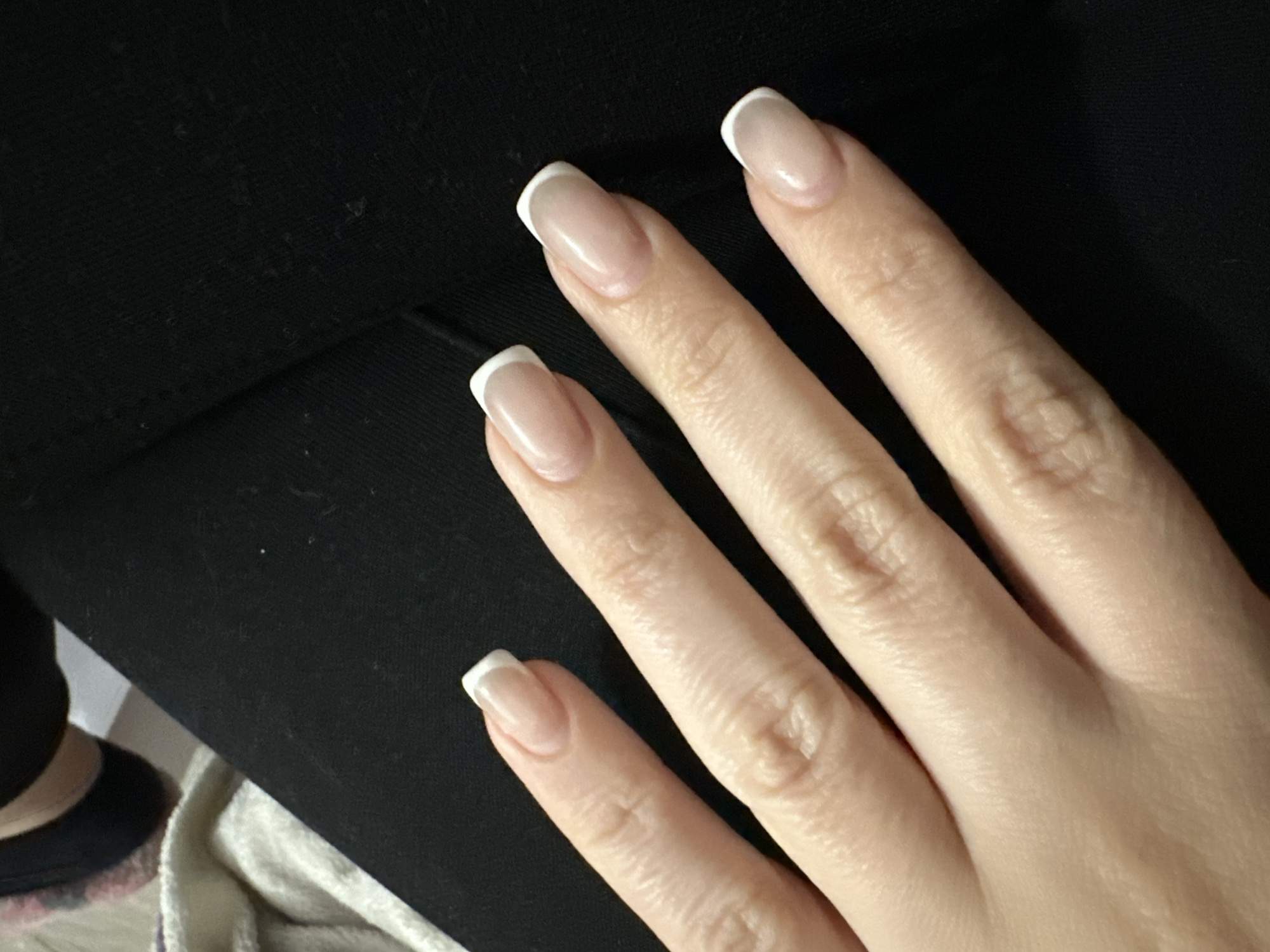 A close-up, high-angle shot features a left human hand with a classic French manicure against a dark background. The hand is fair-skinned, with visible knuckles and faint lines. All five visible fingernails have neatly applied, slightly elongated French tips, featuring a natural pink or nude base and crisp white crescent tips. The nails appear well-maintained and glossy.
The background primarily consists of a textured, matte black fabric, likely clothing such as pants, occupying the upper and left portions of the frame. Small, light-colored specks, possibly dust or lint, are scattered across the dark fabric. In the lower-left corner, a blurry section reveals a partial view of what appears to be a foot or ankle wearing a black item, possibly a sock or shoe cuff, resting beside a soft, cream-colored fabric, which could be a blanket or towel. The lighting suggests an indoor setting, and no specific time of day or outdoor weather conditions can be determined. There is no visible text in the image. The overall impression is a casual, relaxed setting showcasing the manicure.