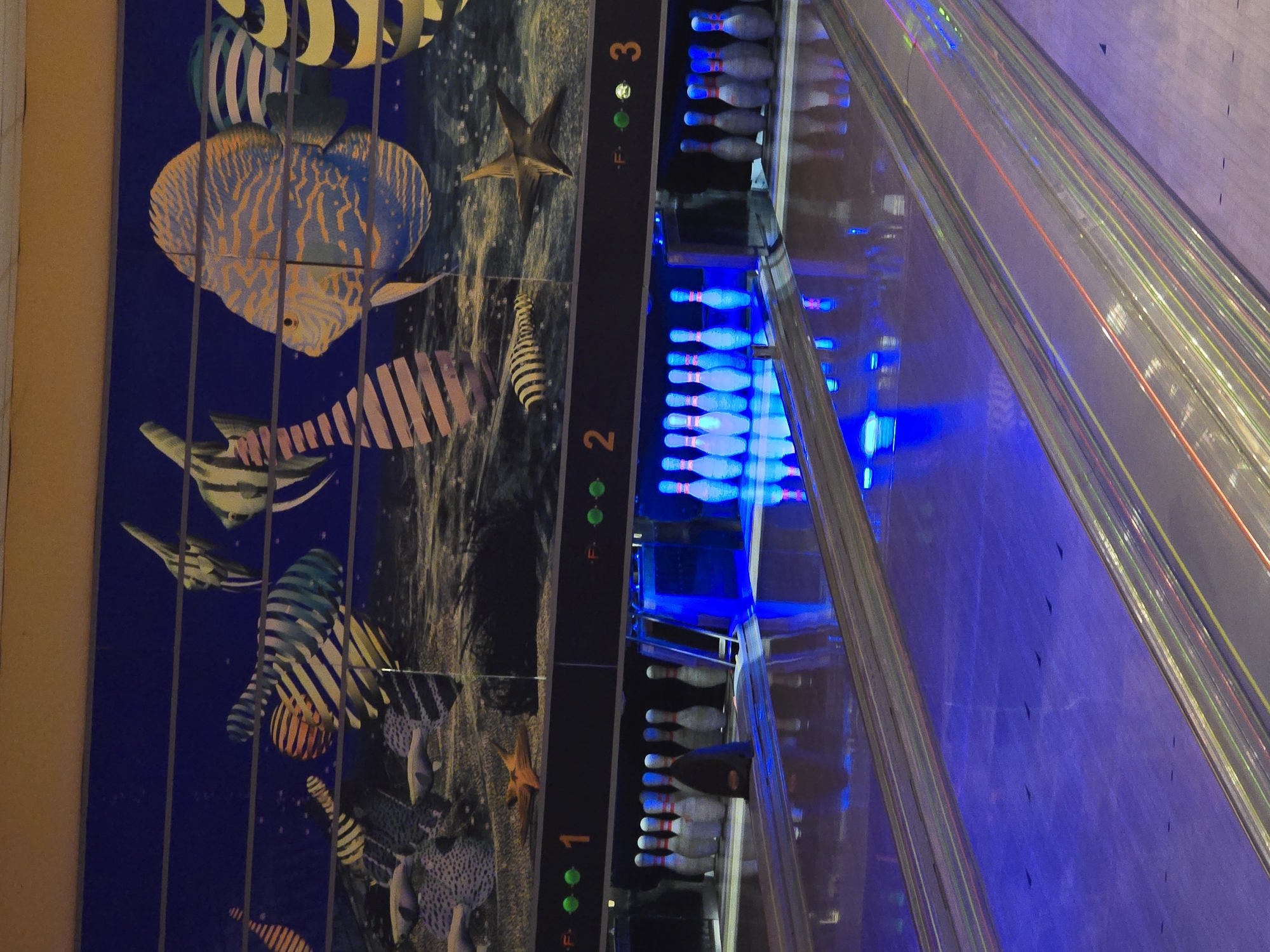 The image depicts the interior of a bowling alley, possibly located in Chaikovsky, Russia. The photograph shows bowling lanes numbered 1, 2, and 3. Each lane contains bowling pins illuminated with blue light. To the left of the lanes is a decorative wall featuring an ocean-themed mural with colorful fish and starfish. The lanes themselves appear to have striped markings on the surface. The overall lighting is dim with strong blue tones. There are no people visible in the frame.