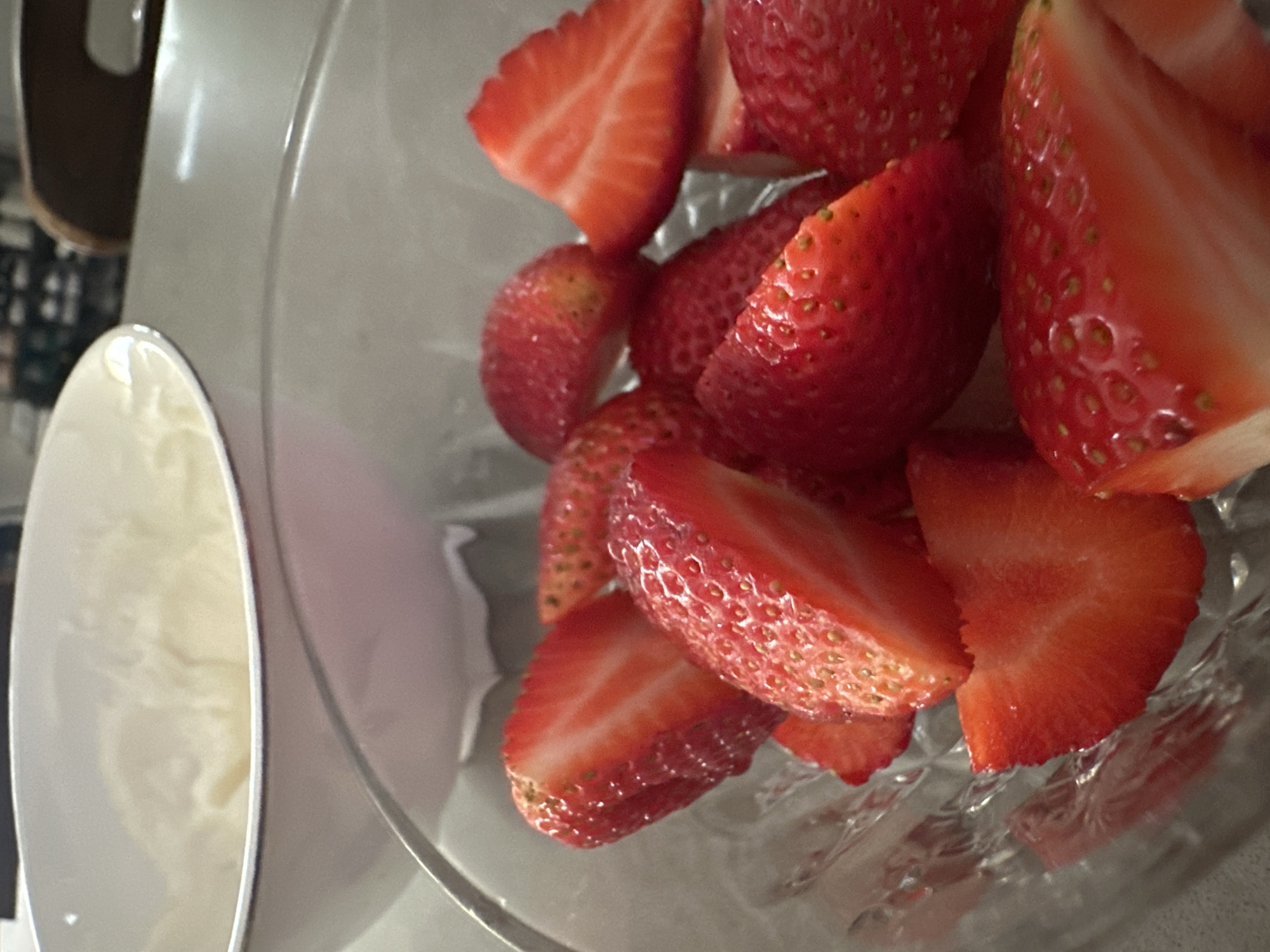 A glass bowl filled with halved and quartered strawberries sits next to a white bowl containing a creamy substance. The scene is located in an indoor setting, possibly a kitchen, in the city of Unknown, Unknown.