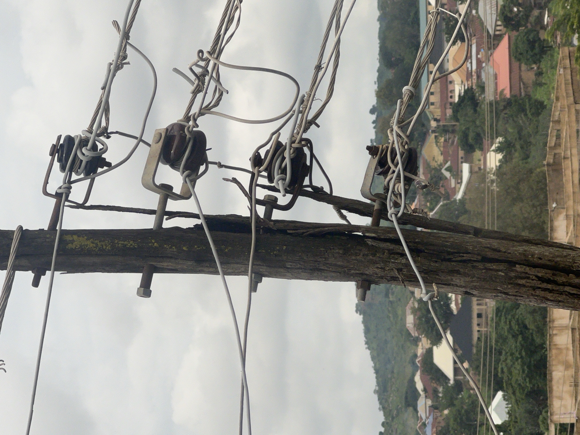 The image captures a weathered wooden utility pole with multiple wires attached, set against a cloudy sky. The pole is centrally located and appears old, with visible cracks and peeling bark. Several ceramic insulators, some dark brown and others a lighter tan, are mounted on metal brackets attached to the pole. These insulators are used to support the numerous electrical wires, which are bundled and intertwined, some appearing taut and others slack. The wires run horizontally across the frame, extending beyond the pole. In the background, a town or village is visible, nestled on rolling green hills. The architecture suggests a warm climate, with buildings featuring a mix of red-tiled and lighter-colored roofs. Dense green trees are scattered throughout the landscape. The overall scene conveys a sense of an established but perhaps not entirely modern infrastructure, typical of many urban or semi-urban areas in Nigeria. The presence of utility poles and overhead wires indicates a functional electrical grid in the depicted settlement, likely within the city of Jos, Nigeria.