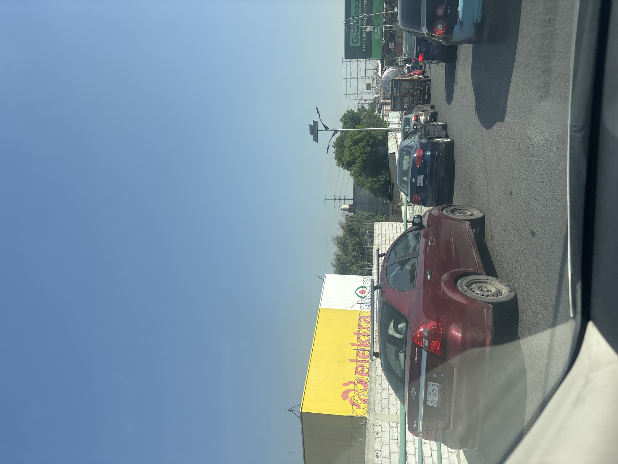 A daylight street scene captures a line of vehicles in what appears to be slow-moving traffic or a jam, viewed from inside a car through a window. The sky is clear and bright blue, indicating good weather.
In the foreground, the rear of a dark red Chevrolet Aveo/Sonic sedan is prominent, showing its license plate "LPX-620-B" with "Puebla" visible below it. The car also has "J.Surman" and "AVEO" badging. Directly behind it are several other cars, including a dark blue Volkswagen sedan with the license plate "PBD-91-84," and a light blue car further down the road. A white truck with what looks like stacked crates is also visible in the traffic line. Shadows of cars are cast on the asphalt road, suggesting the sun is high.
To the left of the road, a large building with a yellow and white facade displays a partially visible logo and the text "Veldekasa". Above the traffic, a green billboard stands tall, featuring the brand name "Grünkleza" and the Spanish slogan "no es suerte es preparación" (it's not luck, it's preparation), along with "Utel". Further in the distance, a street light with mounted solar panels is visible, surrounded by trees and other structures, hinting at an urban or suburban setting in Buenavista, Mexico.
