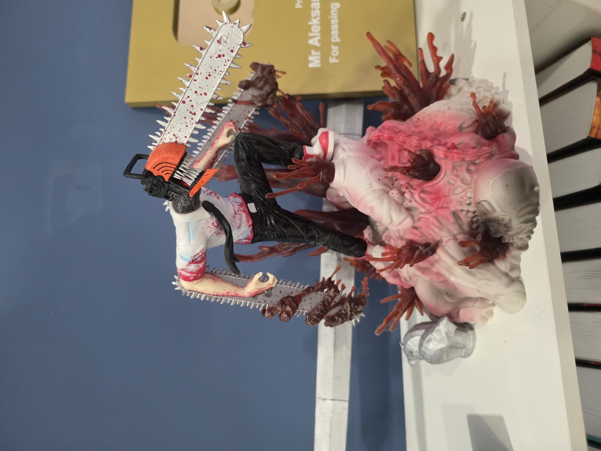 A collectible figurine of the anime character Chainsaw Man, depicted with his signature chainsaw head and a bloody, gore-covered base, is displayed on a white shelf. The figurine is in focus, with a blue wall and a portion of a yellow sign in the background. The scene is indoors, on a shelf that appears to be part of a display in a home or retail setting. There are no people or animals present in the image. The figurine is positioned centrally on the shelf, with other objects out of focus in the background. The lighting is adequate for viewing the figurine clearly. The image does not depict any specific location in the city of Bac Nha Trang Ward, Vietnam; it appears to be a general indoor scene.