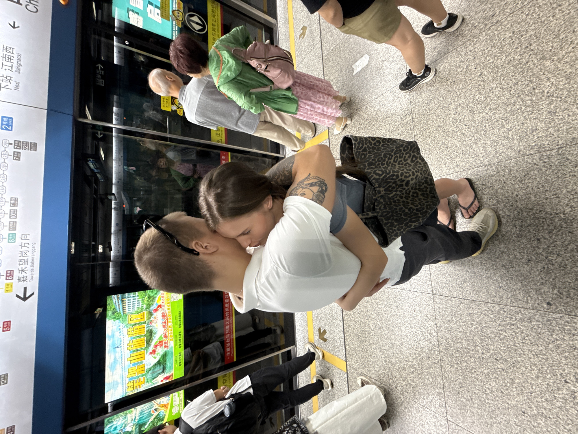 This is a medium-shot, eye-level photograph taken at a subway station in Haizhu District, China, showing a couple embracing in the foreground. The man, wearing a white polo shirt and dark pants, is seen from behind, with his head turned to nuzzle the woman. The woman, also facing away from the camera, is wearing a light blue top and is embraced by the man. She has a large, leopard-print tote bag slung over her left shoulder. Other people are visible in the background, standing and waiting near the subway doors. The subway doors themselves display advertisements. The floor is a speckled gray material with yellow directional lines. Signs in Chinese and English indicating subway routes and destinations are visible above the doors. The overall atmosphere suggests a public transit environment.