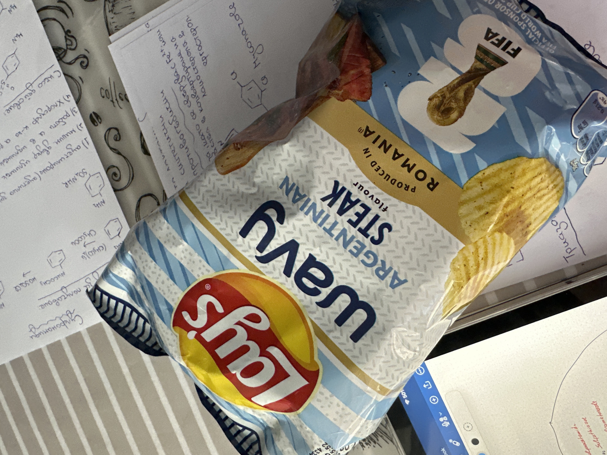 The image shows a close-up, angled view of a study area. Dominating the scene is an open bag of Lay's Wavy Argentinian Steak flavor potato chips, labeled "PRODUCED IN ROMANIA" with the FIFA World Cup logo. Adjacent to the chips are several pages of handwritten notes, seemingly related to organic chemistry, filled with chemical structures and formulas. One page contains the title "Сулфонамиди" (Sulfonamides) written in Cyrillic, and another mentions "Myconazole." The area seems to be a study or work surface, as a laptop screen is visible at the bottom, displaying a document and user interface elements. The environment has a casual, academic feel. Location: The mention of Cyrillic script and chemical notes suggests that the location is Sofia, Bulgaria, due to the presence of Bulgarian notes.