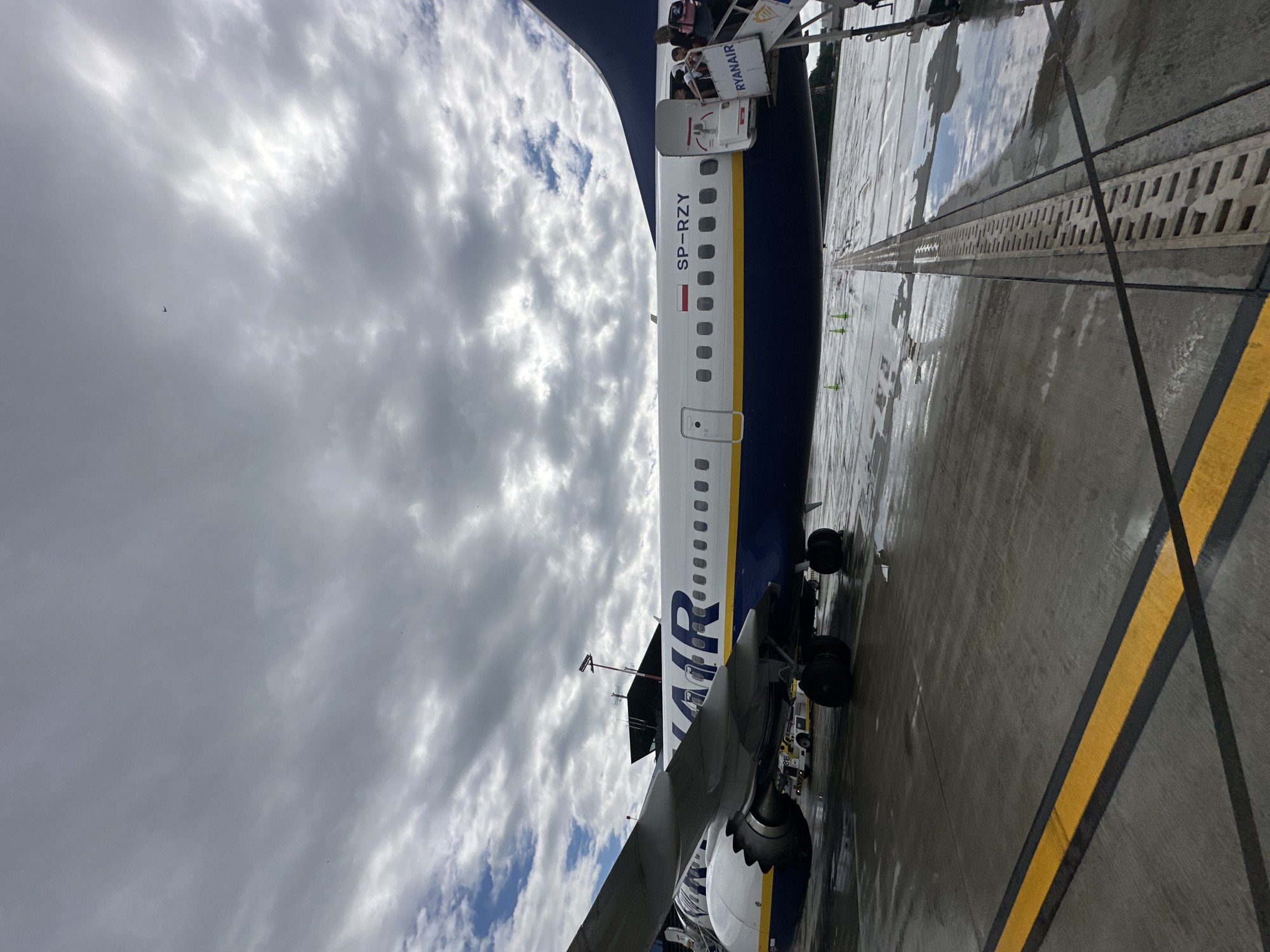 An exterior, eye-level shot shows a Ryanair airplane with the registration SP-RZY parked on a wet tarmac under a cloudy sky at Balice, Poland. The plane is predominantly white with a dark blue stripe along the bottom and the word "RYANAIR" emblazoned in large blue letters. The plane's door is open with a mobile staircase attached, and several passengers are seen ascending the stairs. The tarmac is wet and reflects the cloudy sky. In the background, an airport control tower is visible behind the wing of the airplane. A small baggage tractor is parked near the front landing gear of the aircraft. Yellow and black lines mark the taxiway on the tarmac.