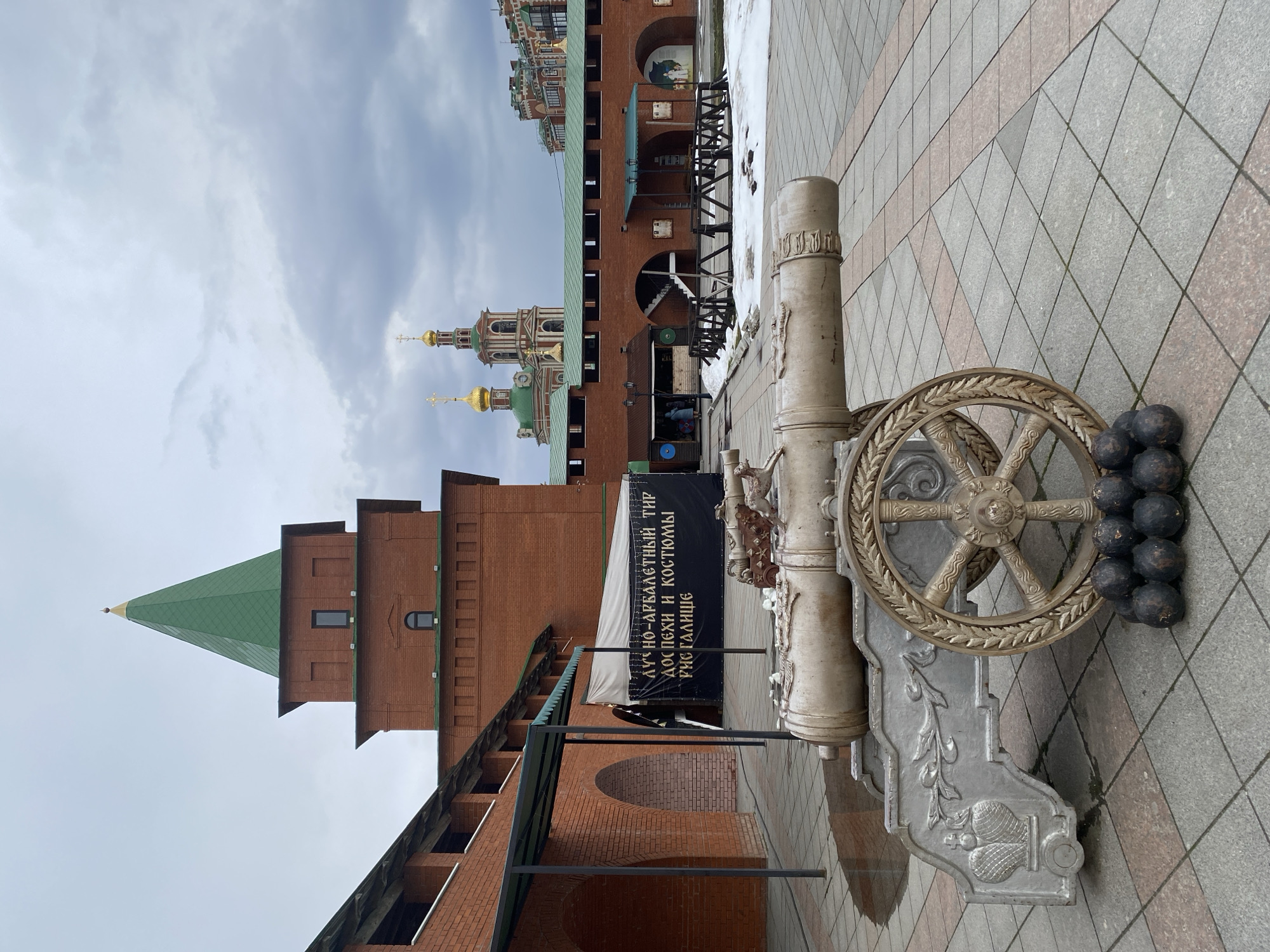 This media file, rotated 90 degrees counter-clockwise, depicts a historical or stylized architectural complex in Yoshkar-Ola, Russia, under an overcast sky. In the foreground, a prominent, light-colored decorative cannon is positioned on a tiled pavement. The cannon features intricate relief carvings on its carriage and a detailed wheel, with several dark cannonballs stacked nearby.
The background features robust red brick buildings. On the left, a tall, multi-tiered tower with a sharp, green conical roof rises towards the sky. Further right, a series of brick walls and structures are visible, some with arched passageways and green-hued roofs. Beyond these, the distinctive golden onion domes of a Russian Orthodox church are prominent against the cloudy sky.
A black banner hangs between two supports, displaying white Cyrillic text. The text reads: "ЛУЧНО-АРБАЛЕТНЫЙ ТИР" (Bow and Crossbow Shooting Range), "ДОСПЕХИ И КОСТЮМЫ" (Armor and Costumes), and "РИСТАЛИЩЕ" (Tournament Grounds/Lists). This indicates a recreational activity or attraction.
Patches of snow are visible on the ground in certain areas, suggesting cold weather, likely late winter or early spring. The pavement consists of light-colored and reddish-brown square tiles arranged in a diagonal pattern. The overall scene conveys a blend of historical architecture and recreational offerings in a public square or courtyard setting. No people are visible.