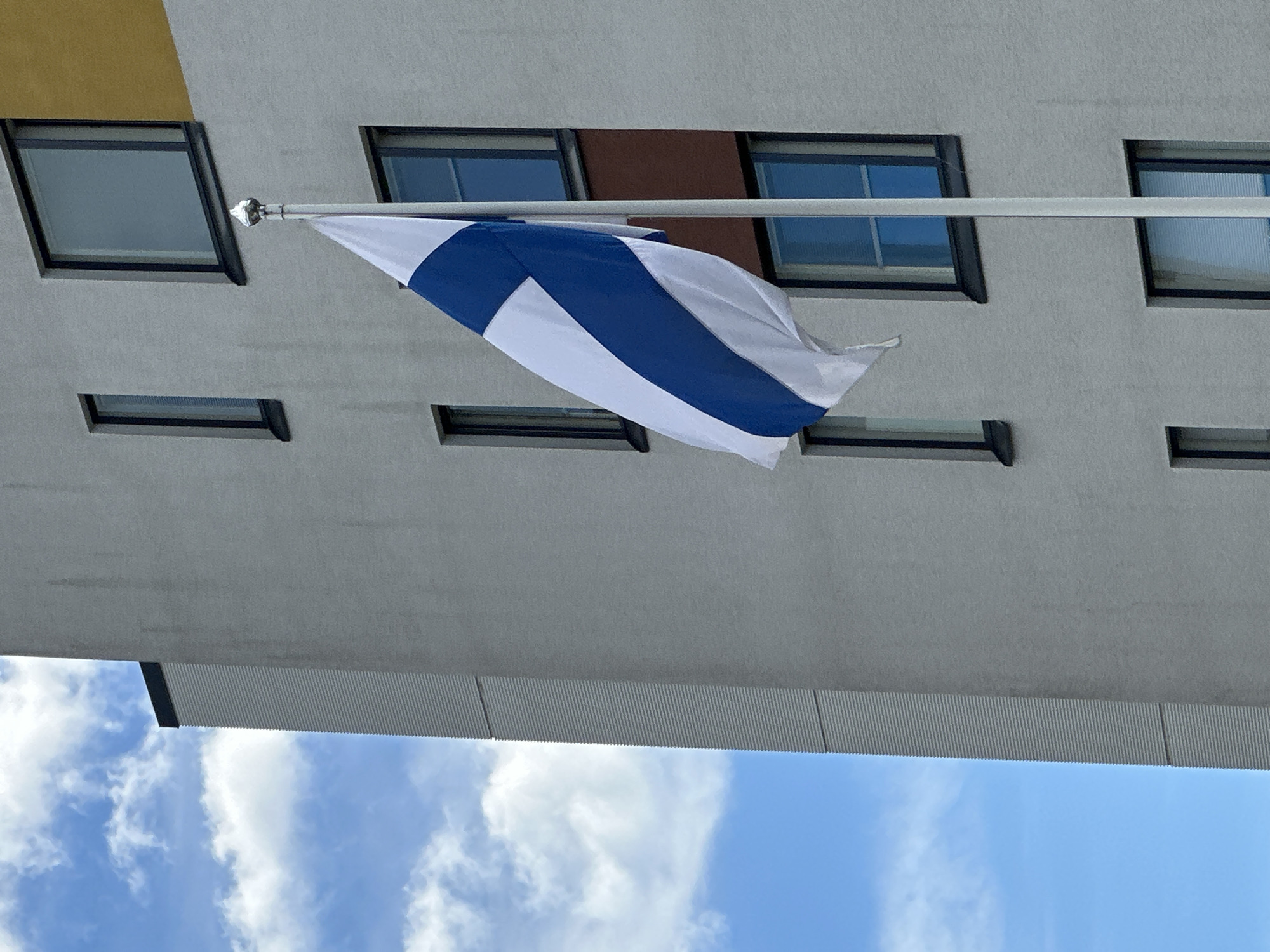 A Finnish flag, white with a blue cross, waves on a flagpole in front of a modern building with a grey facade and yellow and brown accents. The flag is partially obscuring several rectangular windows with dark frames. The sky, visible to the left of the building, is blue with scattered white clouds. A section of the building's facade on the left has a textured, ribbed design.