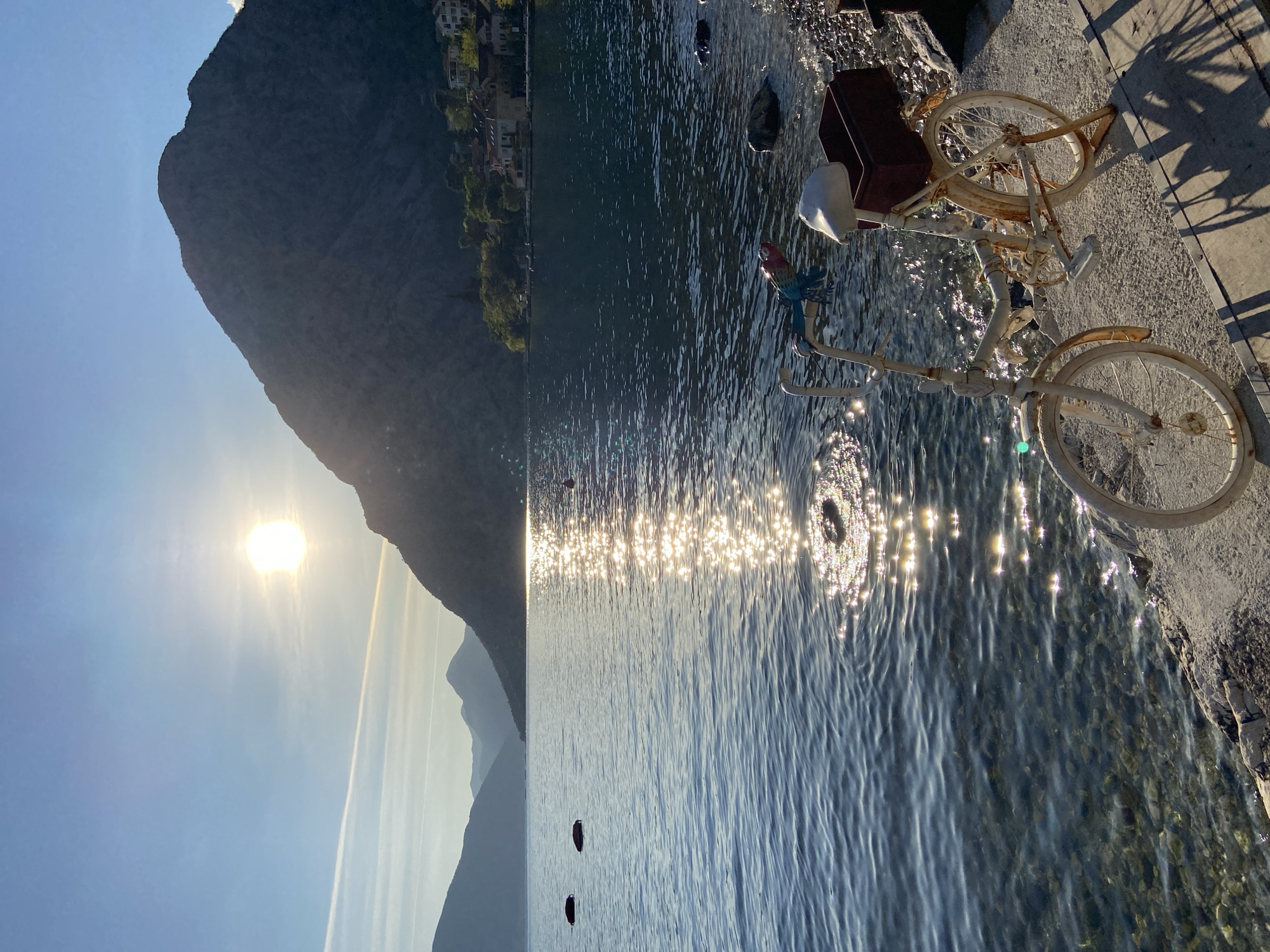 The image shows a sunny day at Donji Orahovac, Montenegro, next to the Bay of Kotor. A tall mountain rises on the left of the image. The sun is positioned high above the mountain. The water in the bay is shimmering with reflections of the sun. Two black round objects appear on the surface of the water. On the right is a concrete platform next to the water. A weathered, white bicycle is parked on the platform, near the water's edge. It has a brown basket behind the seat. A small, colorful decoration hangs from the bike near the water. A rock sits in the water beside the bike. Buildings can be seen at the base of the mountain across the bay.
