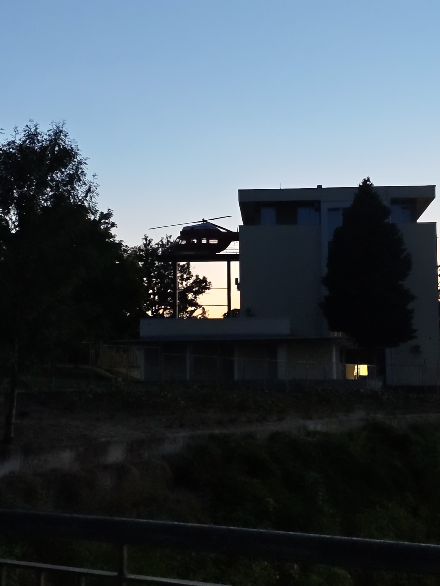 The image depicts a building with a helicopter on its roof, possibly a helipad, against a clear sky. The building appears to be modern with large windows. The surrounding area is silhouetted against the twilight sky, showing trees and a landscape that could be interpreted as a park or a natural setting adjacent to an urban area. The presence of a helicopter on a building suggests a helipad, which could be located at a hospital, police station, or a private facility. Given the context of Pleven, Bulgaria, this could be the helipad of the Pleven Regional Hospital, which is known to have one for medical transport. The image captures a moment of transition from day to night, with the last rays of sunlight reflecting on the windows. The overall atmosphere is serene, yet tinged with the potential for urgent activity given the helicopter's presence.
