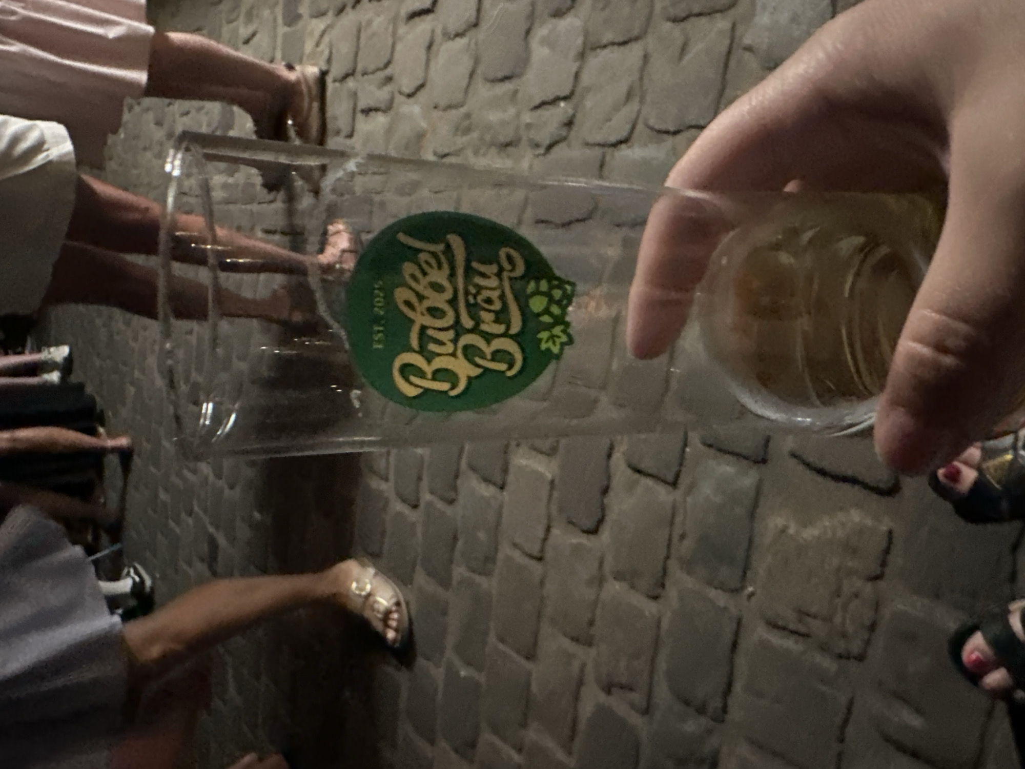 In Krefeld, Germany, on a cobblestone street, a person's hand holds a nearly empty glass beer mug with a green circular logo featuring the text "Bubbel Bräu" and "EST. 2025" in yellow. The mug has a small amount of amber liquid at the bottom. In the background, blurred figures of people are visible, wearing sandals and dresses. The lighting suggests it is evening or nighttime.